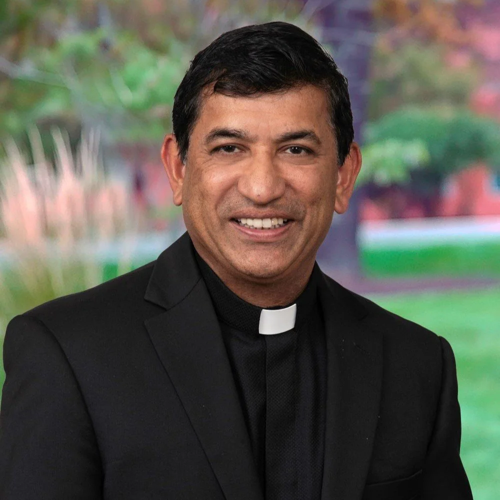 Headshot of a smiling man wearing a black clerical shirt with a white clerical collar, against a gradient grey background.