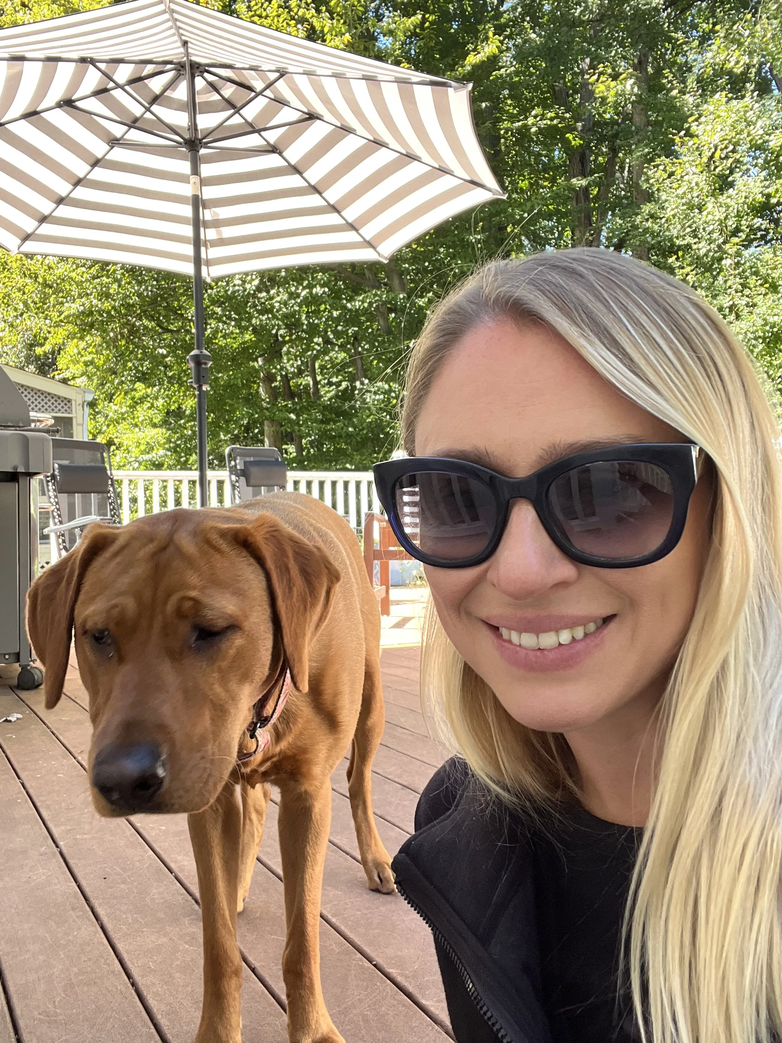 A woman with blonde hair and sunglasses smiling next to a brown dog on a wooden deck outdoors, with a large striped patio umbrella and green trees in the background.