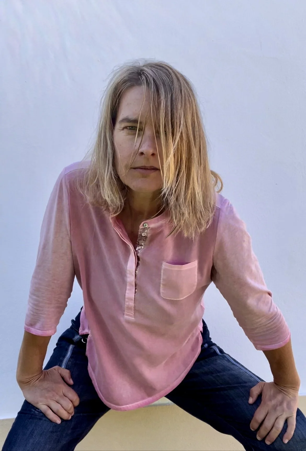 Woman in a pink long-sleeve shirt and dark jeans squatting with hands on knees, looking into the camera with half her face partially covered by her hair, against a plain white wall background.