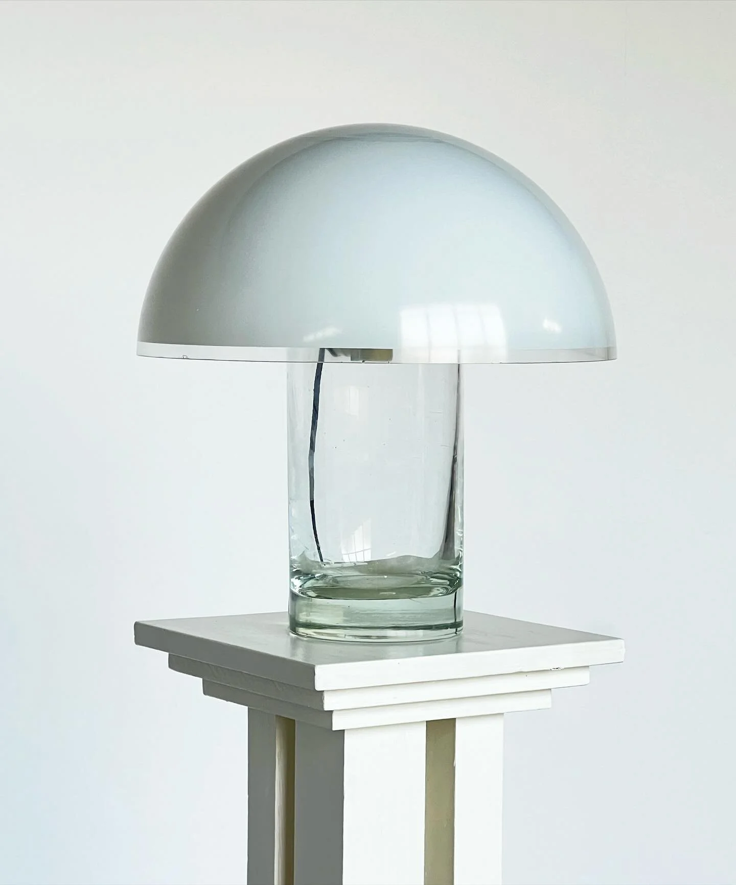 SOLD A minimal Italian table lamp with a cylindrical clear glass base and a wide domed white glass shade. Its clean geometry and contrasting materials create a striking silhouette, characteristic of 1970s modernist Italian design. Both functional and