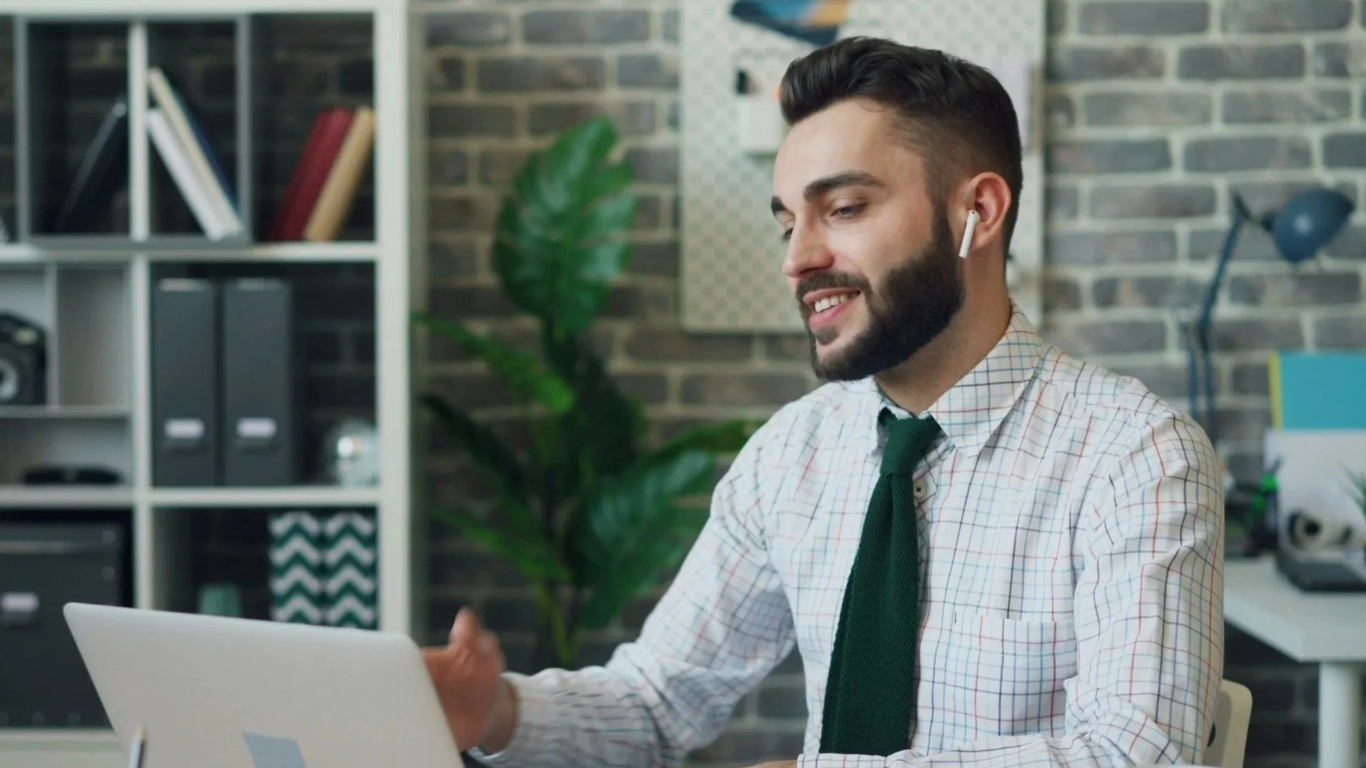 A man with dark hair and a beard, wearing a checkered shirt and green tie, smiling while wearing wireless earbuds, sitting at a desk with a laptop in an office setting.