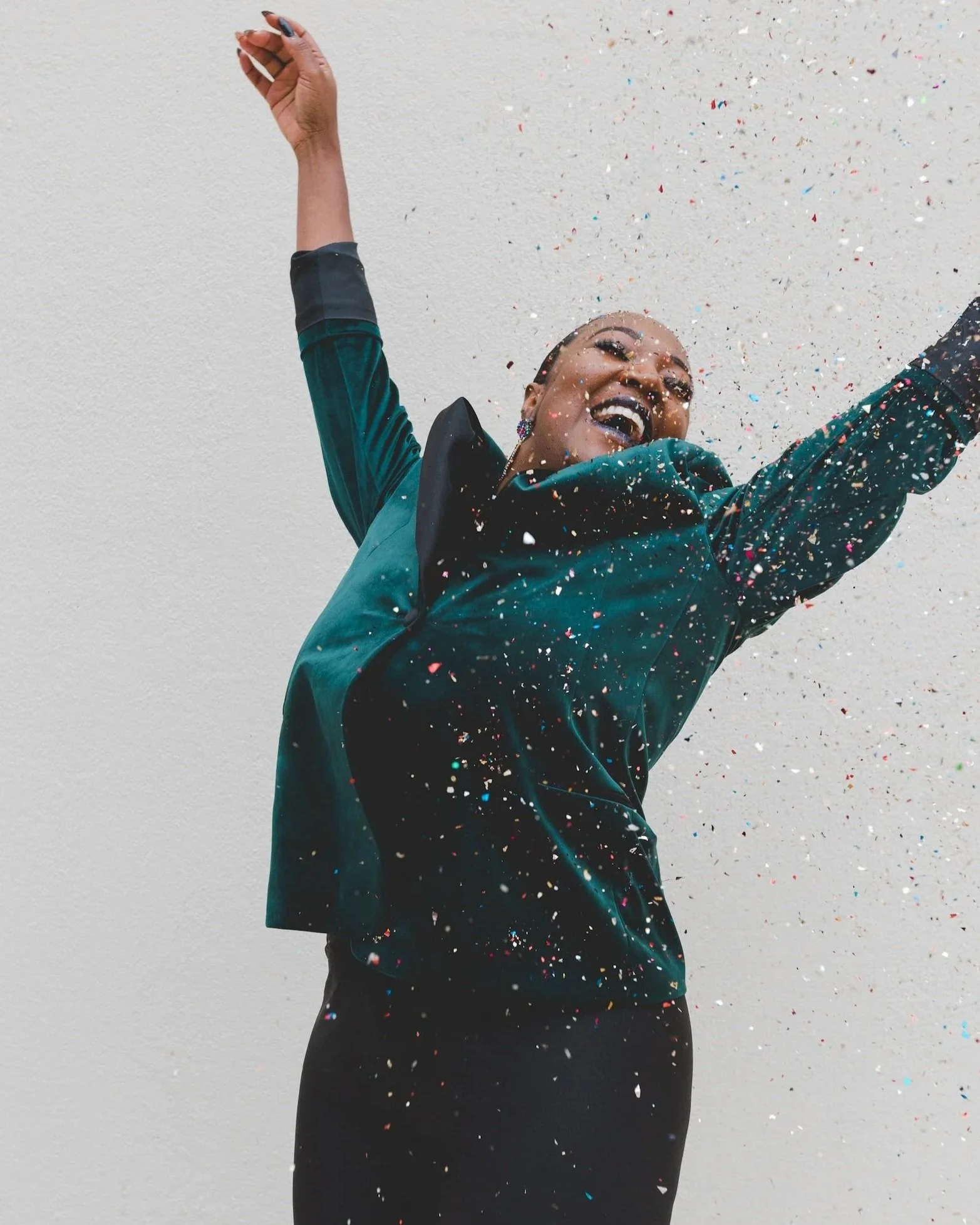 A woman joyfully celebrating with arms raised, surrounded by colorful confetti against a plain white wall.