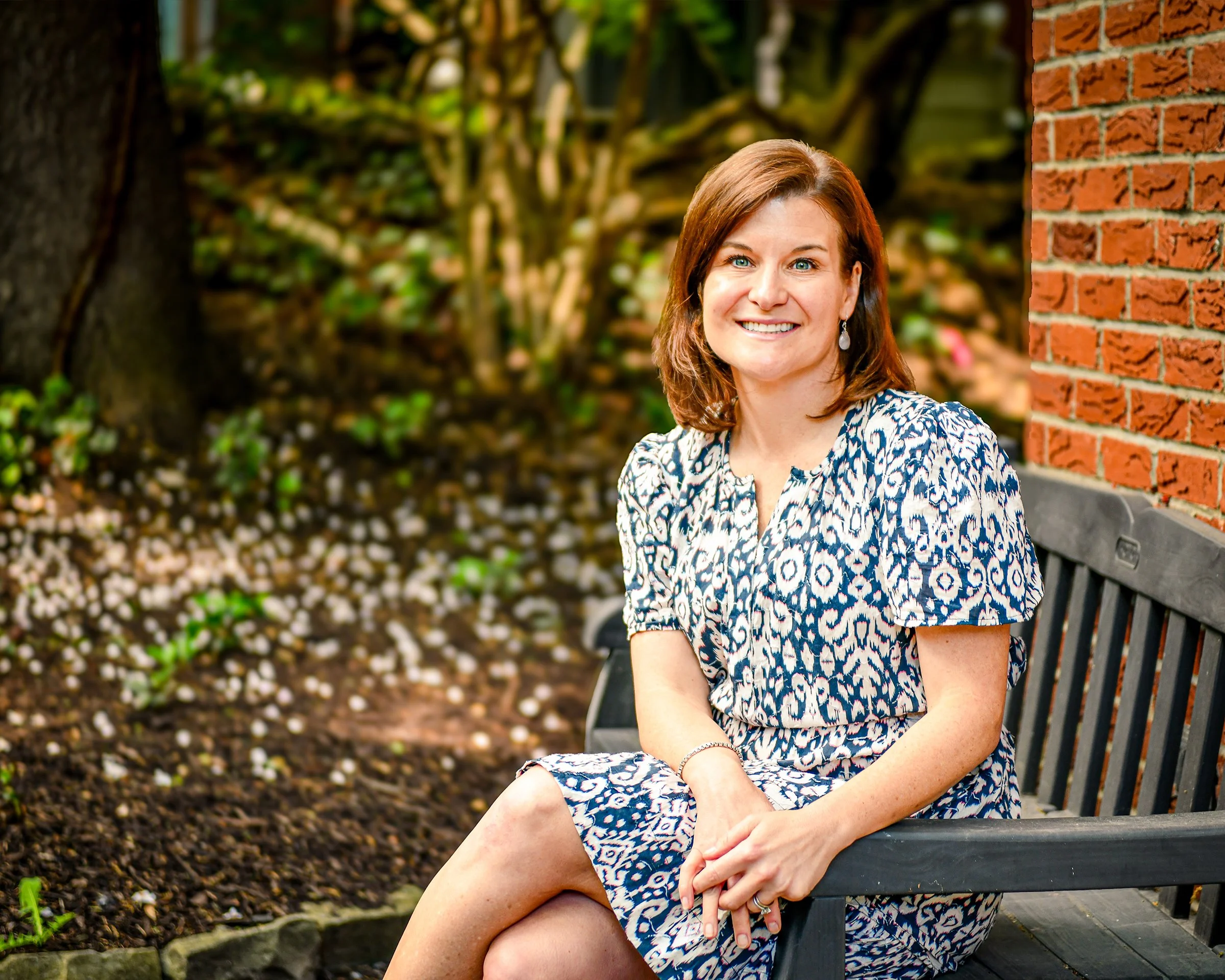 A woman with shoulder-length reddish-brown hair and blue eyes sitting on a wooden bench outdoors near a brick wall, smiling at the camera, wearing a patterned blue and white dress and earrings, with greenery and a large tree in the background.