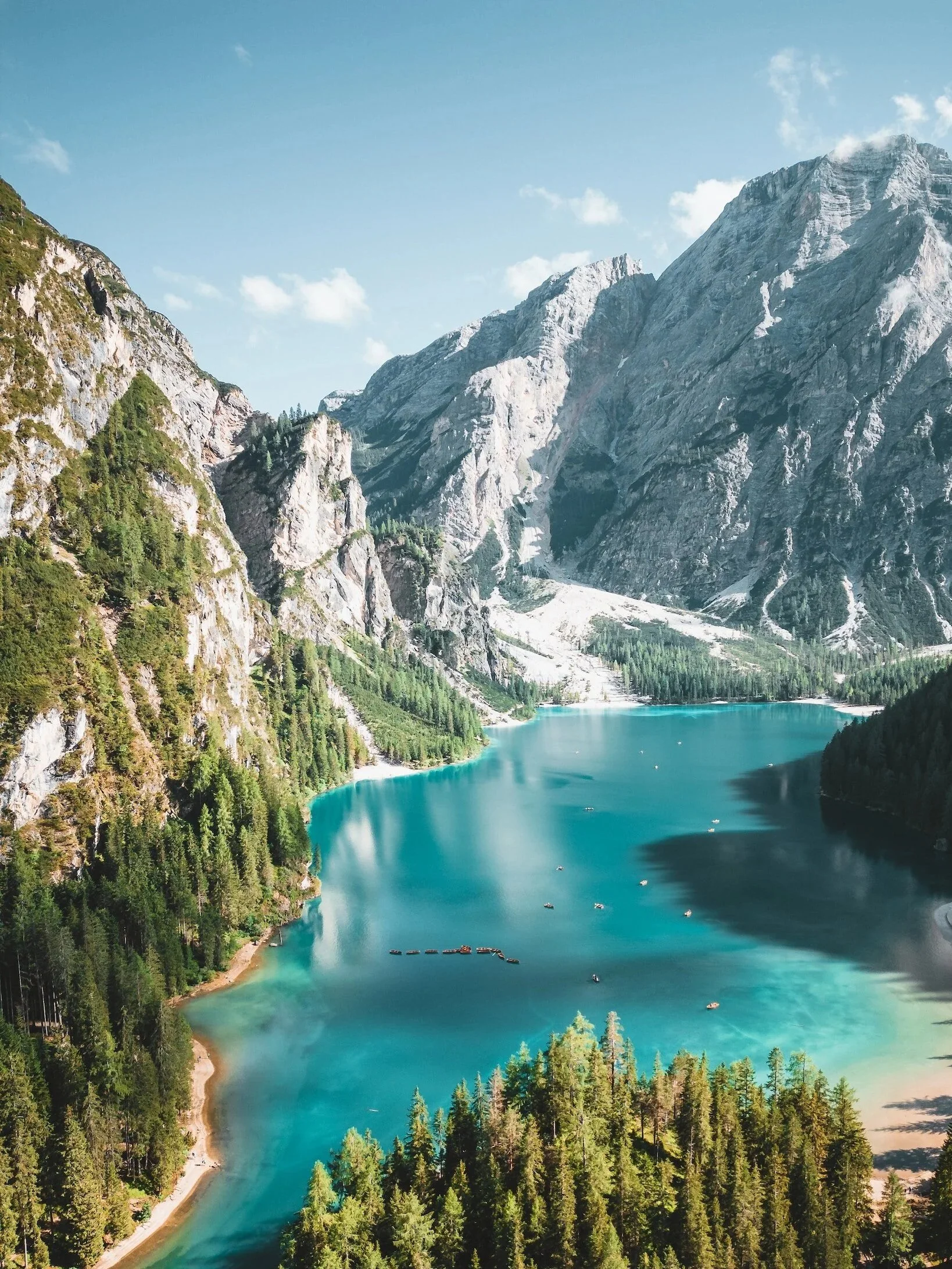 A scenic view of a turquoise lake surrounded by steep mountains with rocky cliffs and lush green forests under a partly cloudy sky.