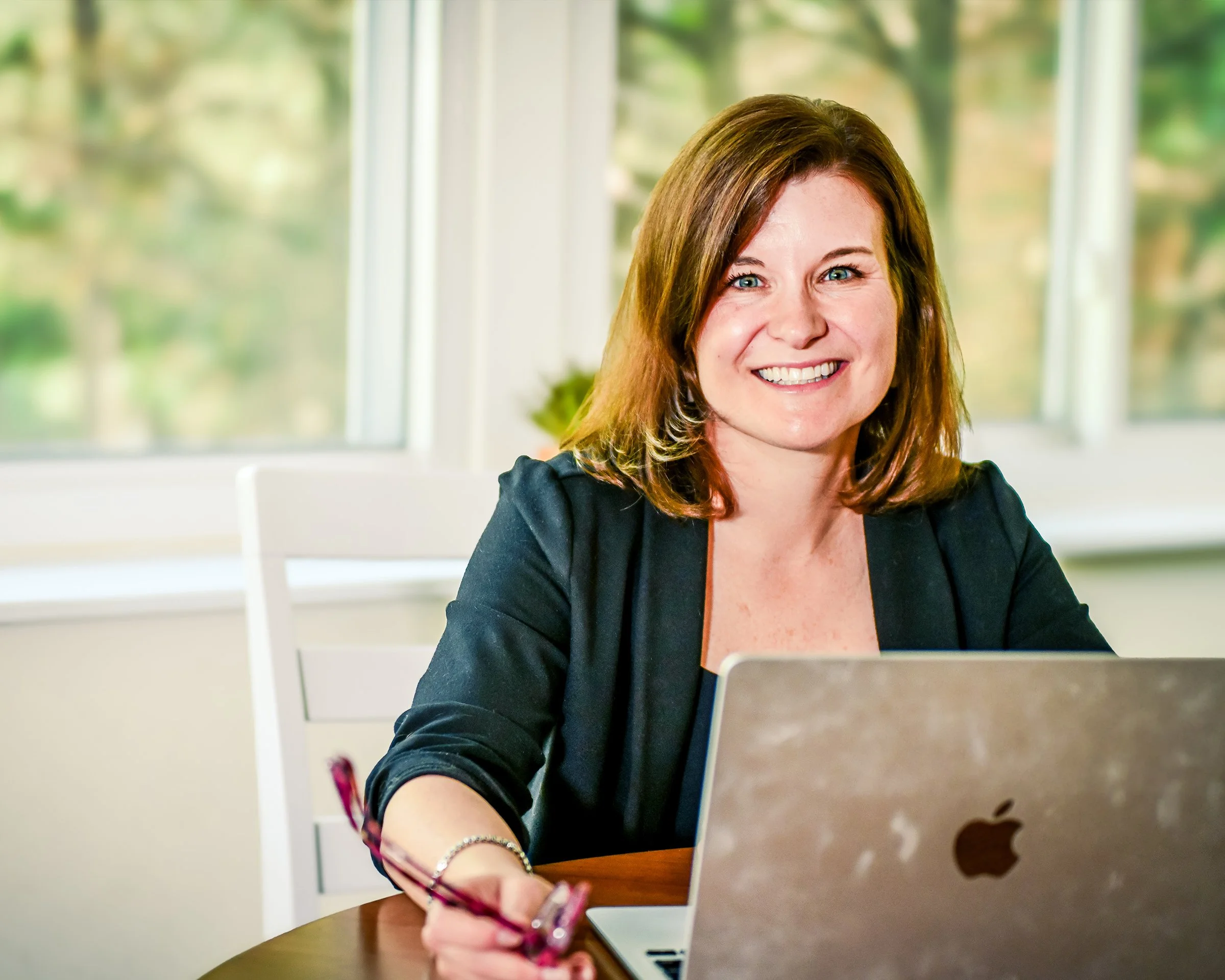 A woman with auburn hair smiling while working on a laptop at a table in a bright room with large windows and green trees outside.