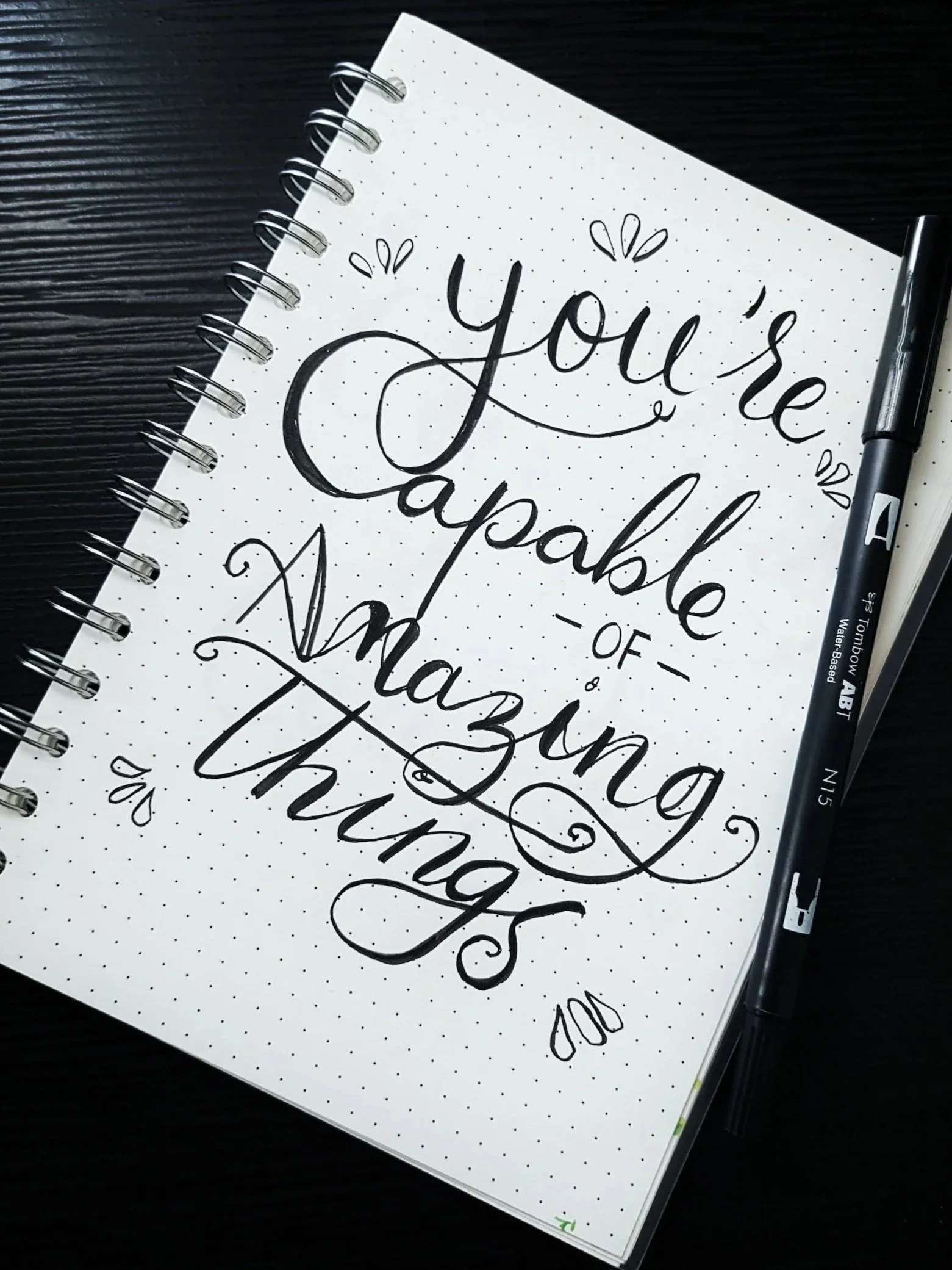 A spiral-bound notepad with handwritten calligraphy that reads 'You're capable of amazing things', placed on a dark wooden surface. A black marker lies beside the notepad.