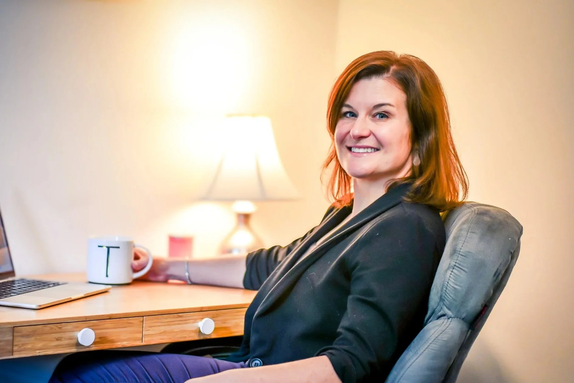 A woman with reddish-brown hair smiling and sitting in a gray armchair. She is wearing a black blazer and is holding a white mug with a letter T on it. There is a wooden desk with a laptop and a smartphone in front of her. A table lamp and a wall are in the background.