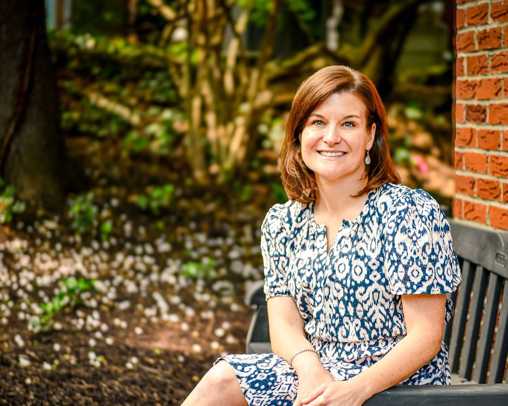 A woman with shoulder-length brown hair and blue eyes sitting on a black bench outdoors next to a brick wall, smiling, with green trees and foliage in the background.