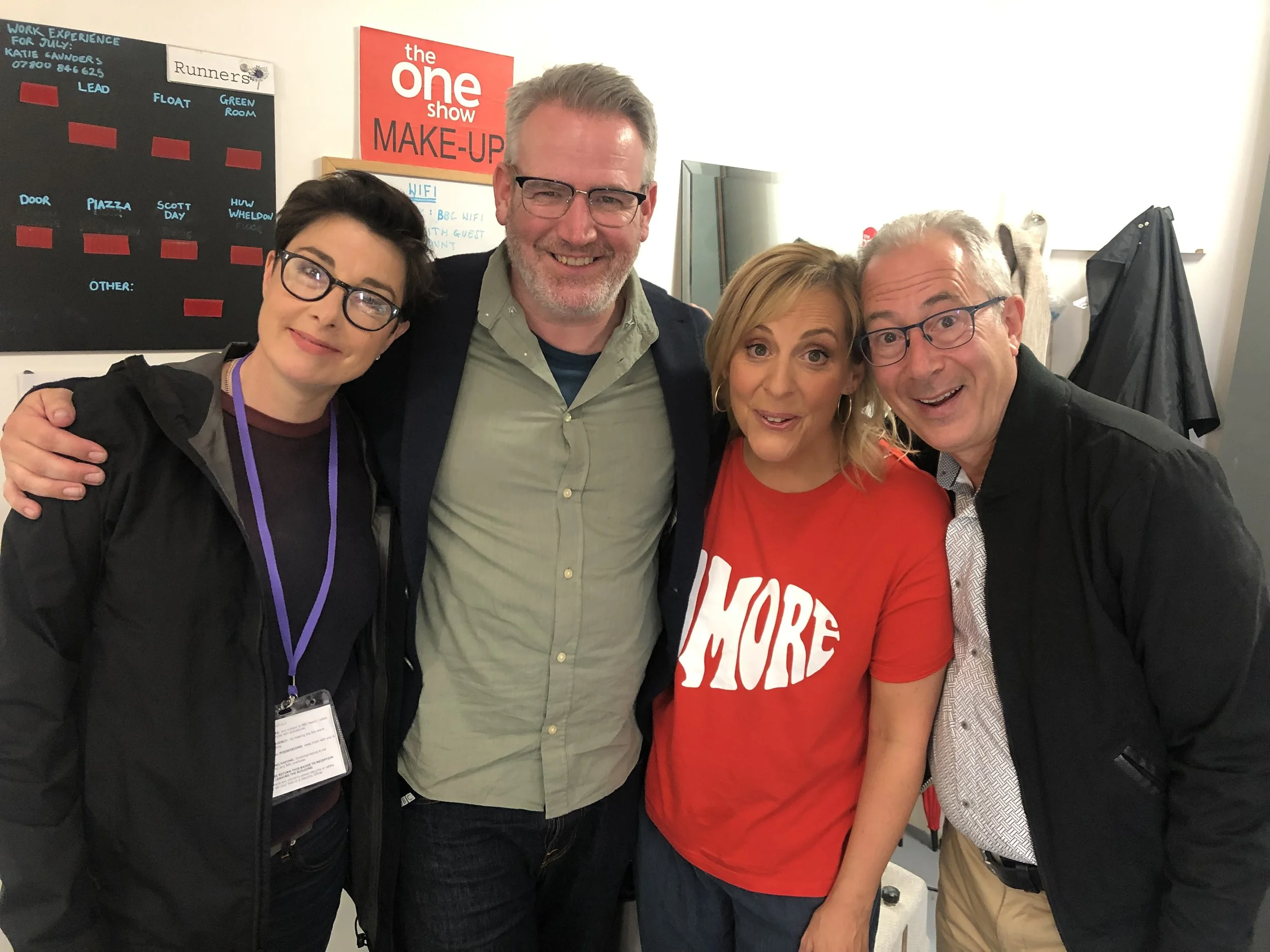 Group of four individuals smiling, standing close together indoors next to a wall with a sign saying "The One Show Make-Up." The background includes a black chart and a whiteboard with text and notes.