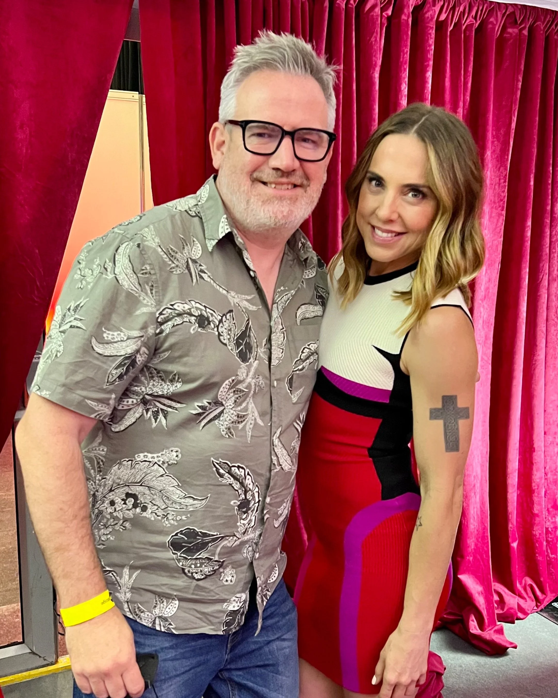 Two people posing in front of red curtains, both smiling. The person on the left is wearing glasses and a patterned shirt, while the person on the right is wearing a sleeveless dress and has a tattoo on their arm.