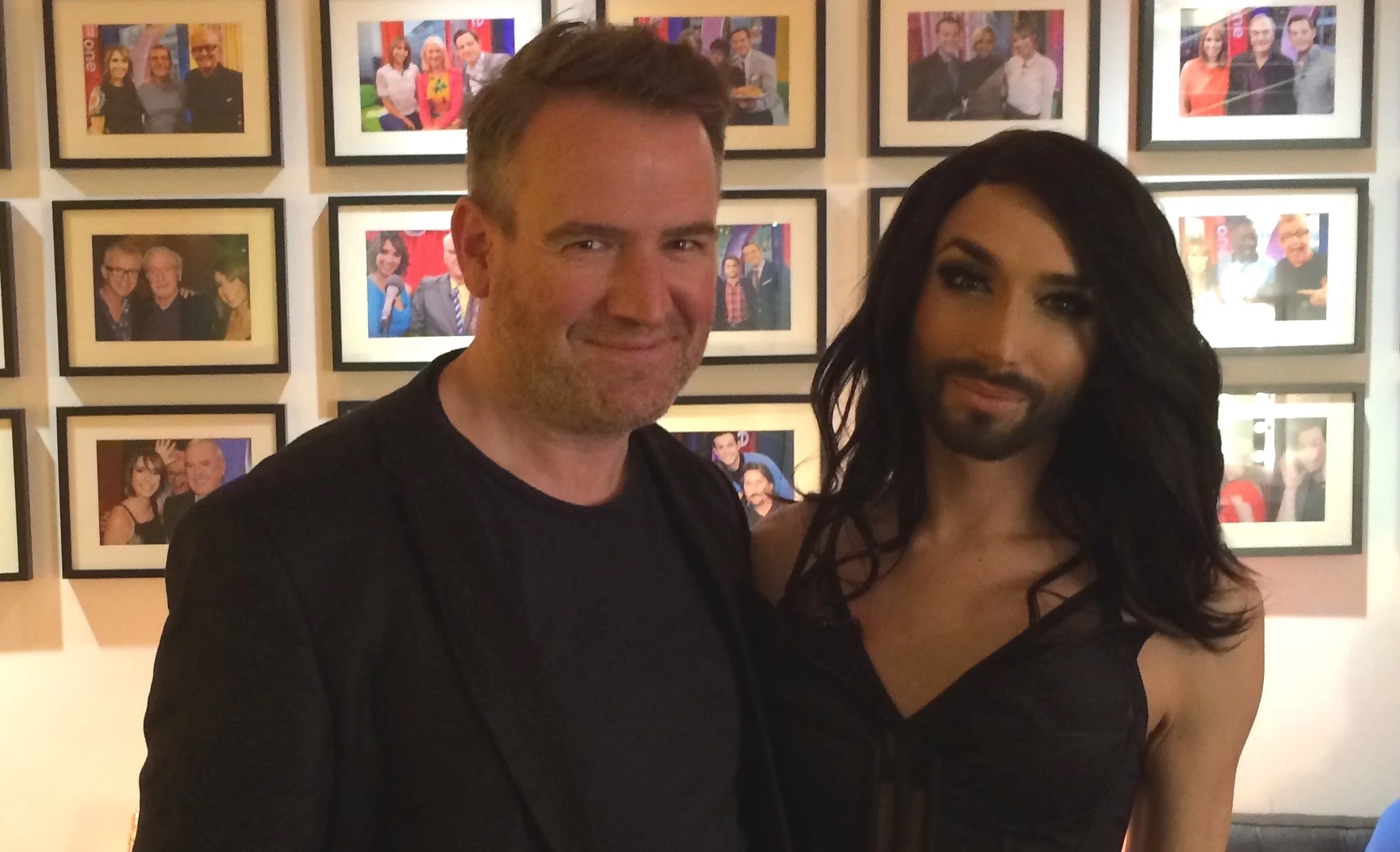 Two people posing in front of a wall with framed photos, in a brightly lit room.
