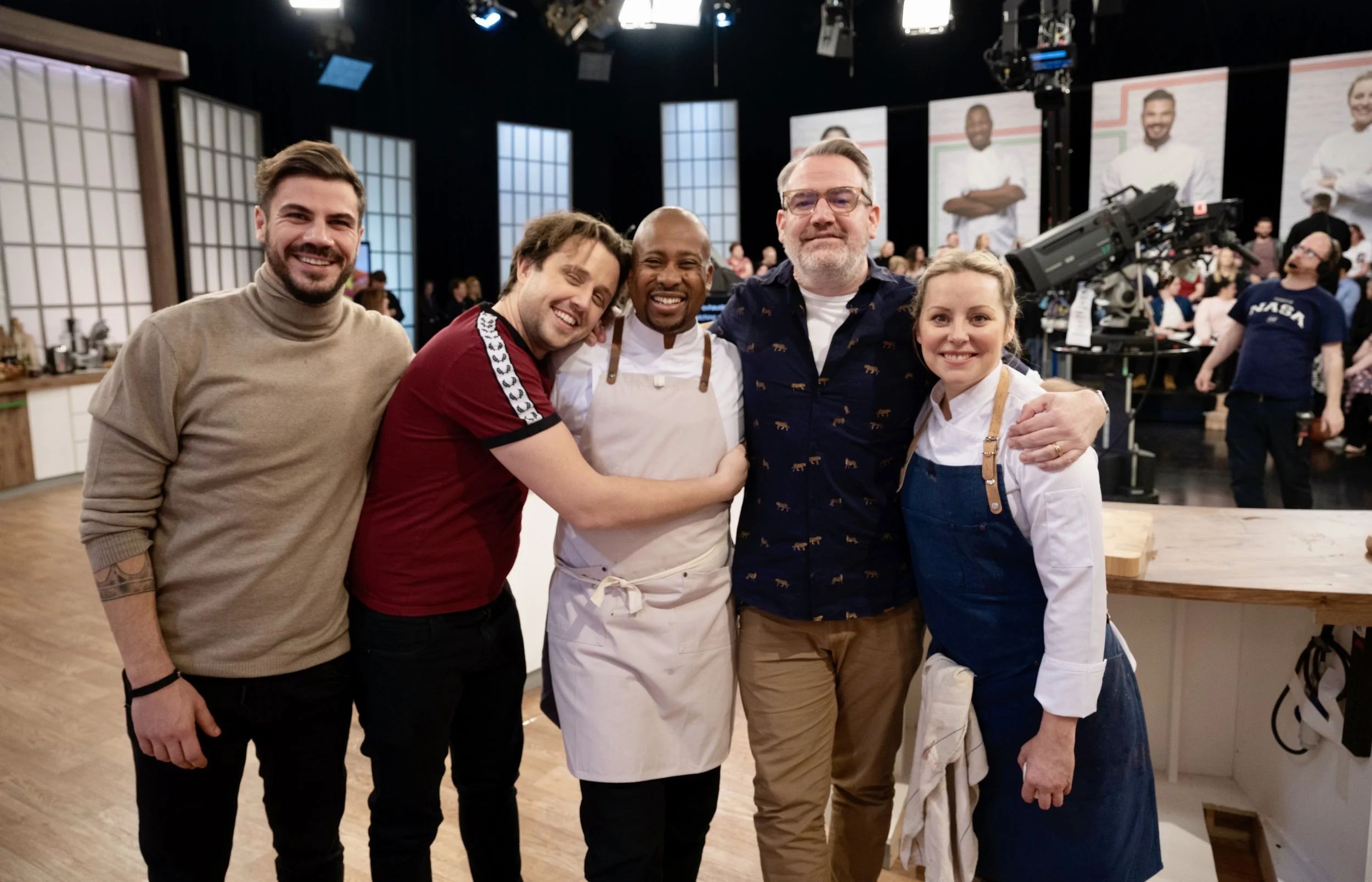 Group of five people smiling and hugging in a studio kitchen setting, with cooking equipment and cameras in the background.