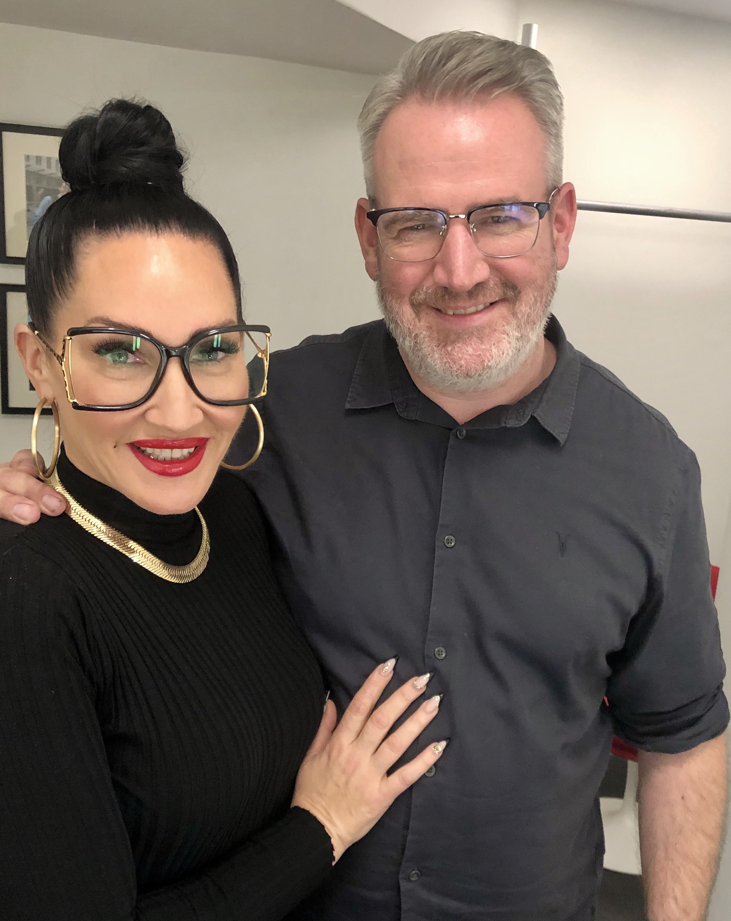 Two people smiling, one with glasses and a bun, wearing gold jewelry, and the other wearing glasses and a black shirt.