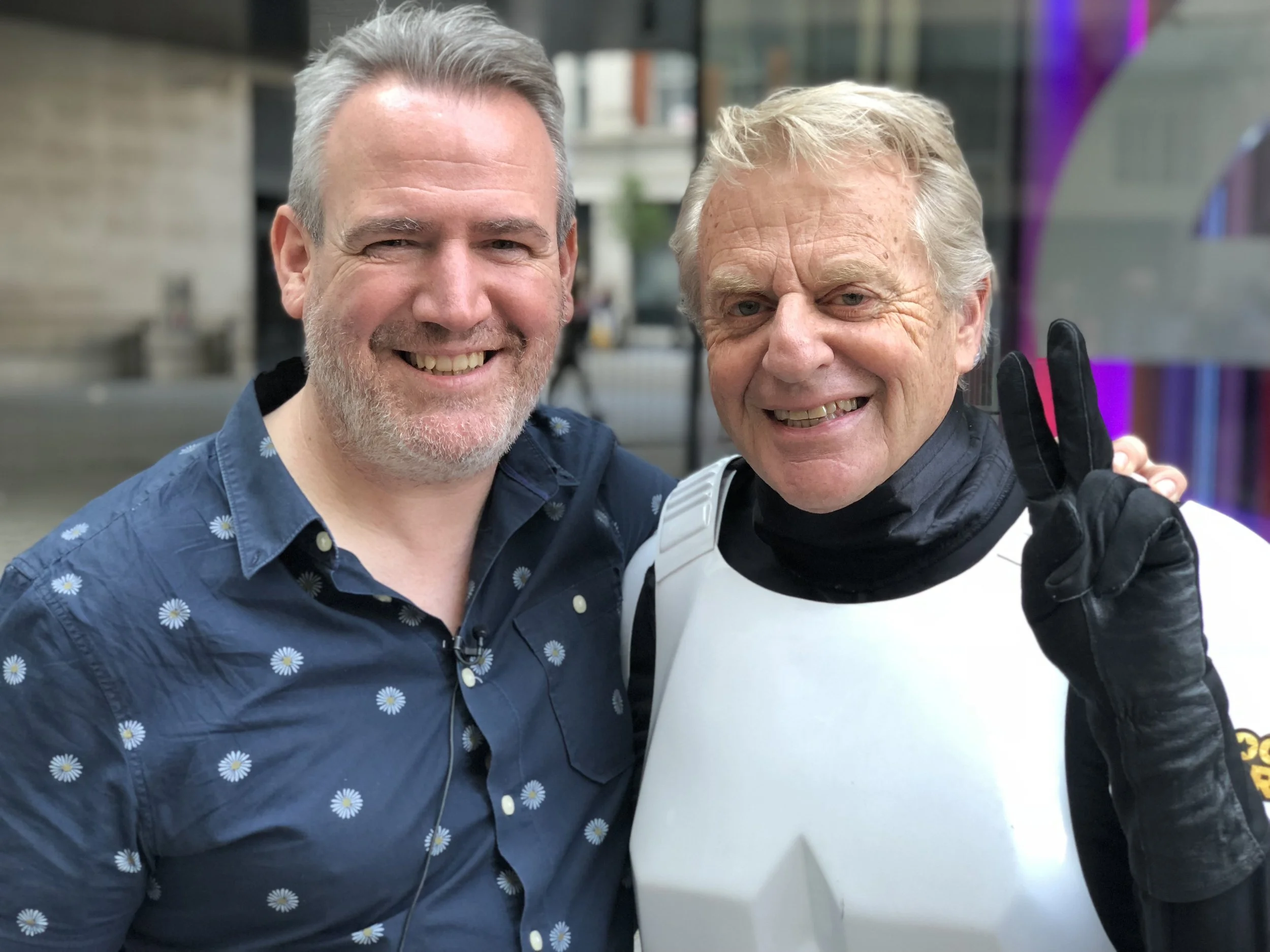 Two smiling men posing; one wearing a floral shirt, the other in white armor with gloves giving a peace sign.