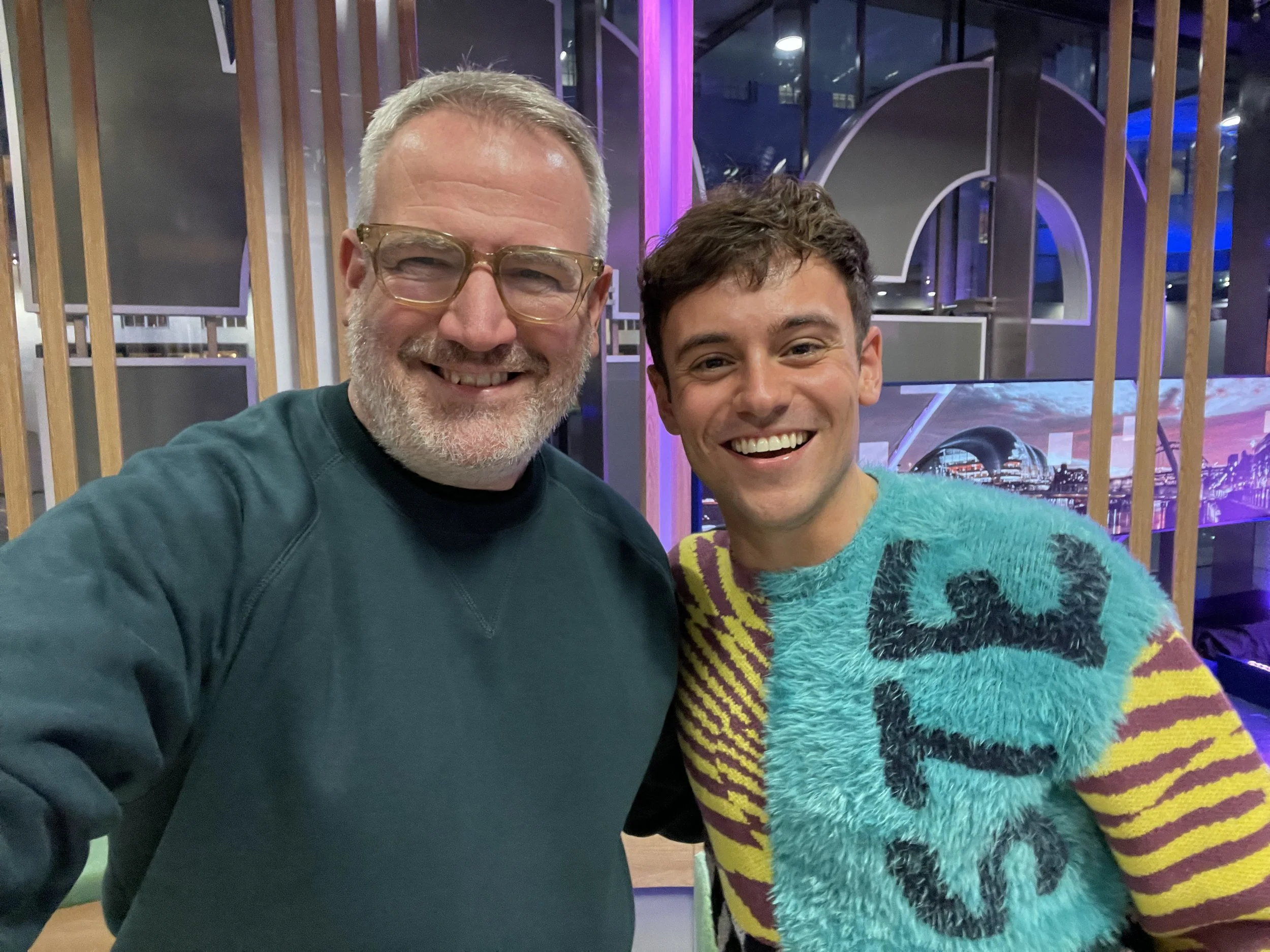 Two men smiling indoors, one wearing glasses and a teal sweater, the other in a colorful fuzzy sweater with "ST5" partially visible. Background includes wooden accents and a screen with an urban scene.