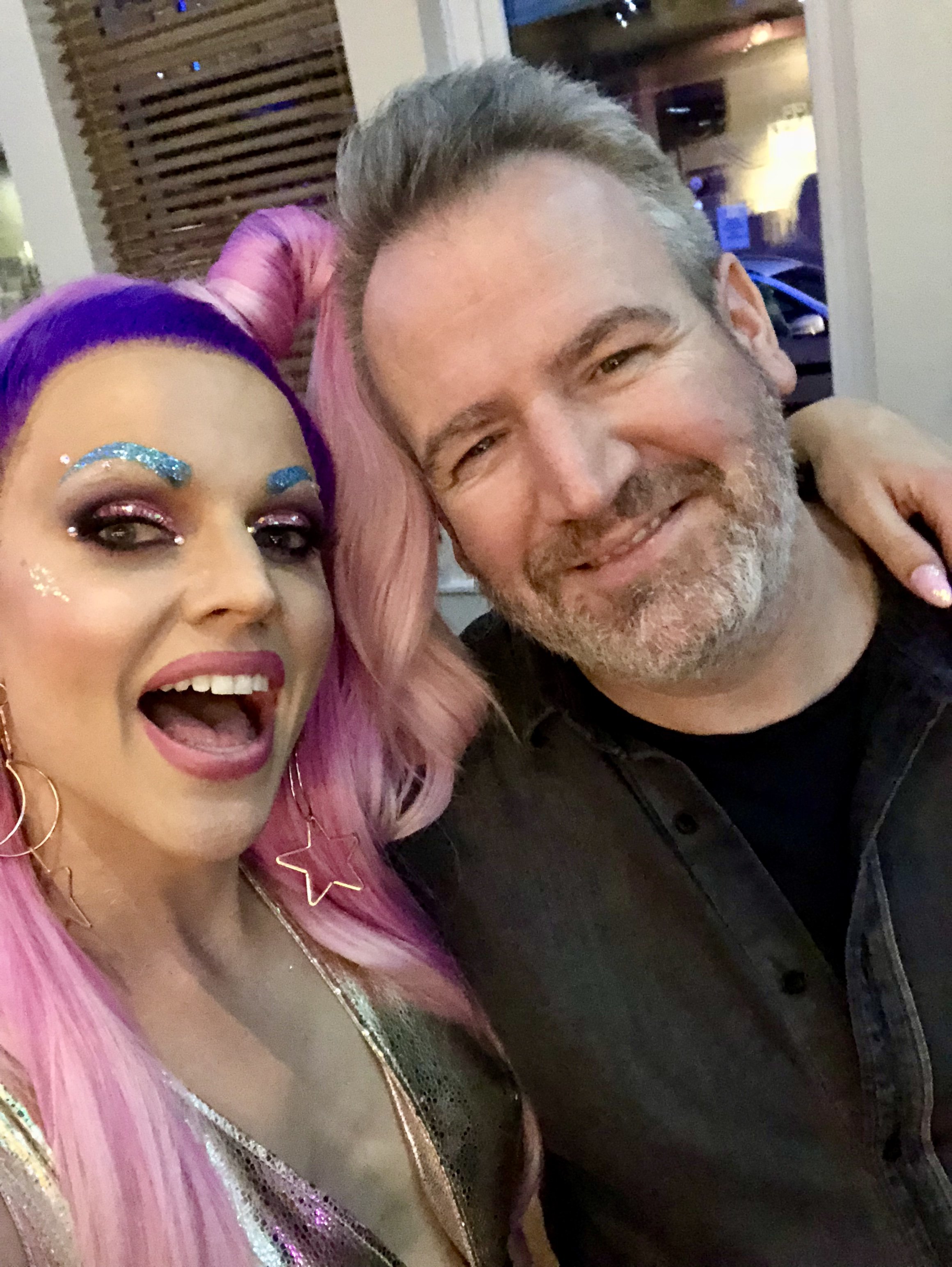 Two individuals posing for a selfie; one with purple and pink hair, colorful makeup, and star earrings, the other with short gray hair and a beard, both smiling.