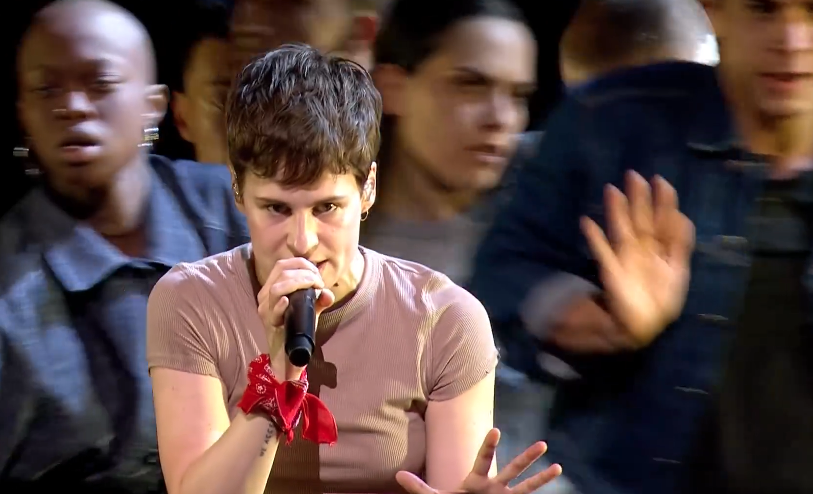 Person singing on stage with microphone, wearing red bandana and surrounded by blurred dancers.