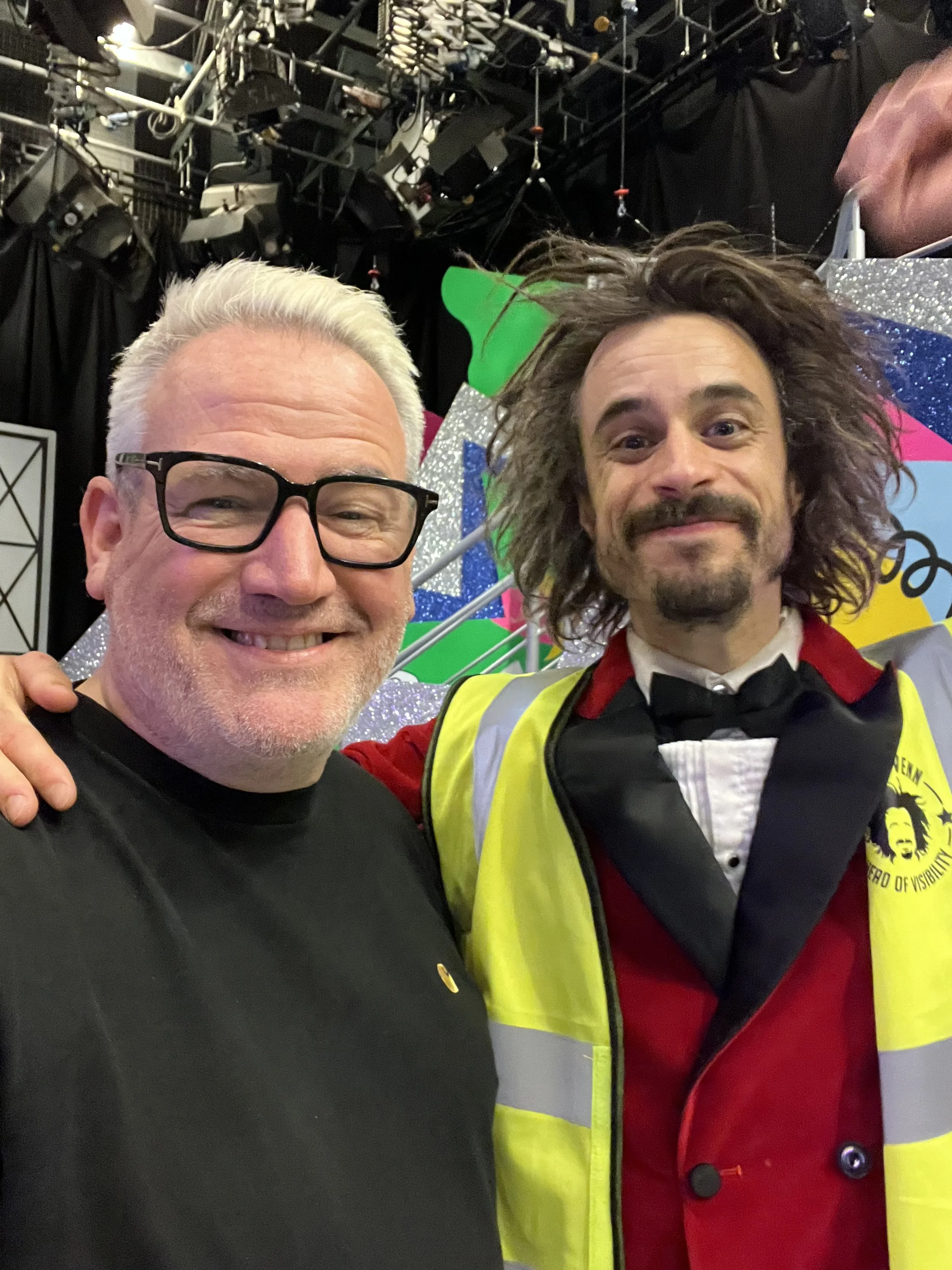 Two people smiling indoors, one wearing glasses and a black shirt, the other in a red jacket with a reflective vest. Bright, decorated set background with stage lighting overhead.