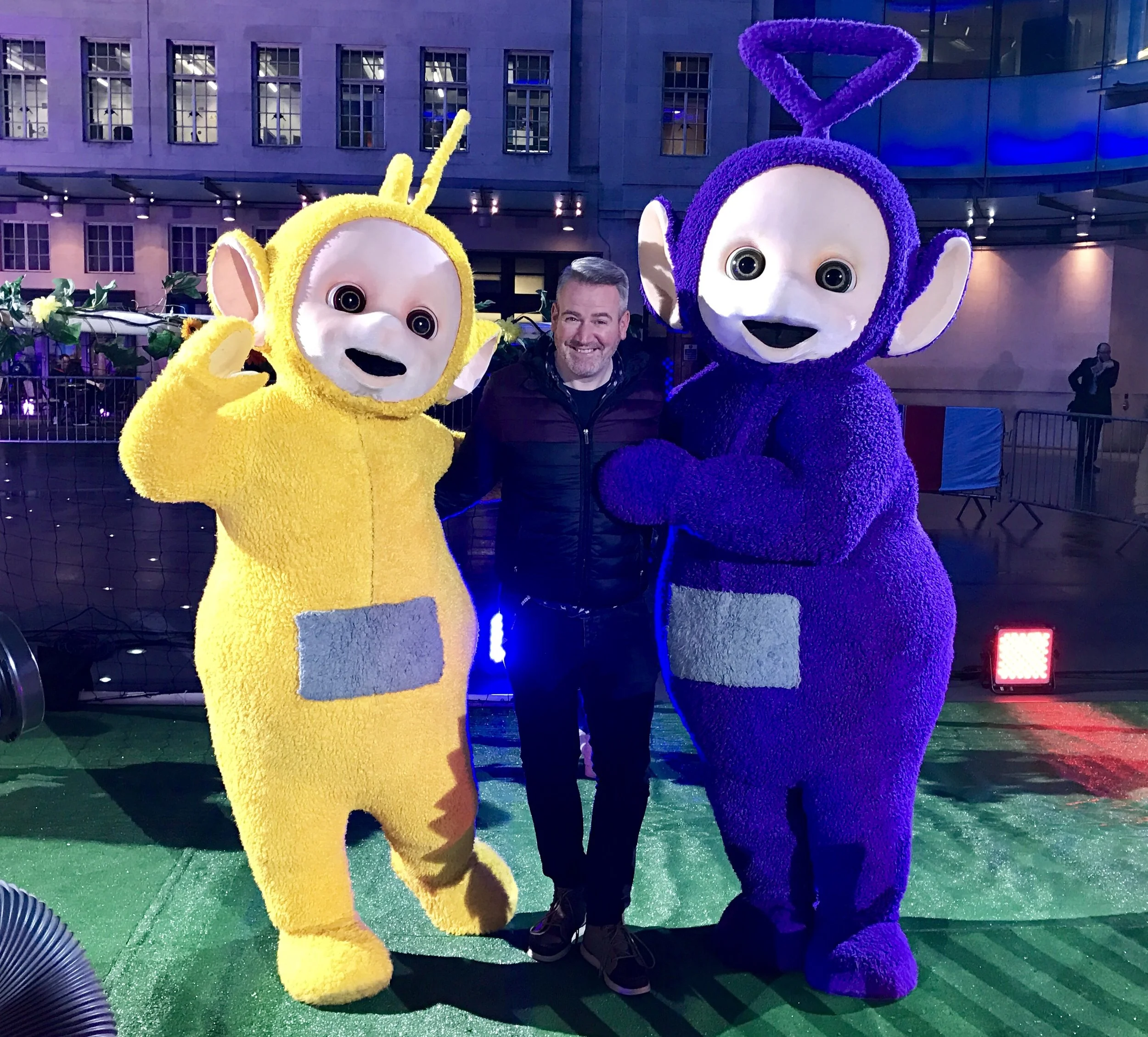Person posing with two Teletubby mascots, one yellow and one purple, outdoors at night.