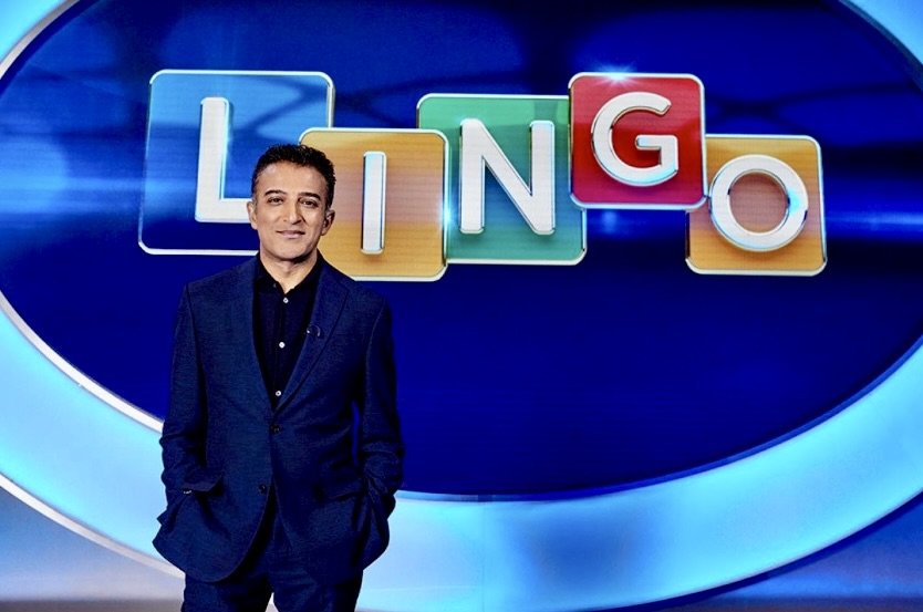 Person standing in front of colorful letters spelling "LINGO" on a game show set.