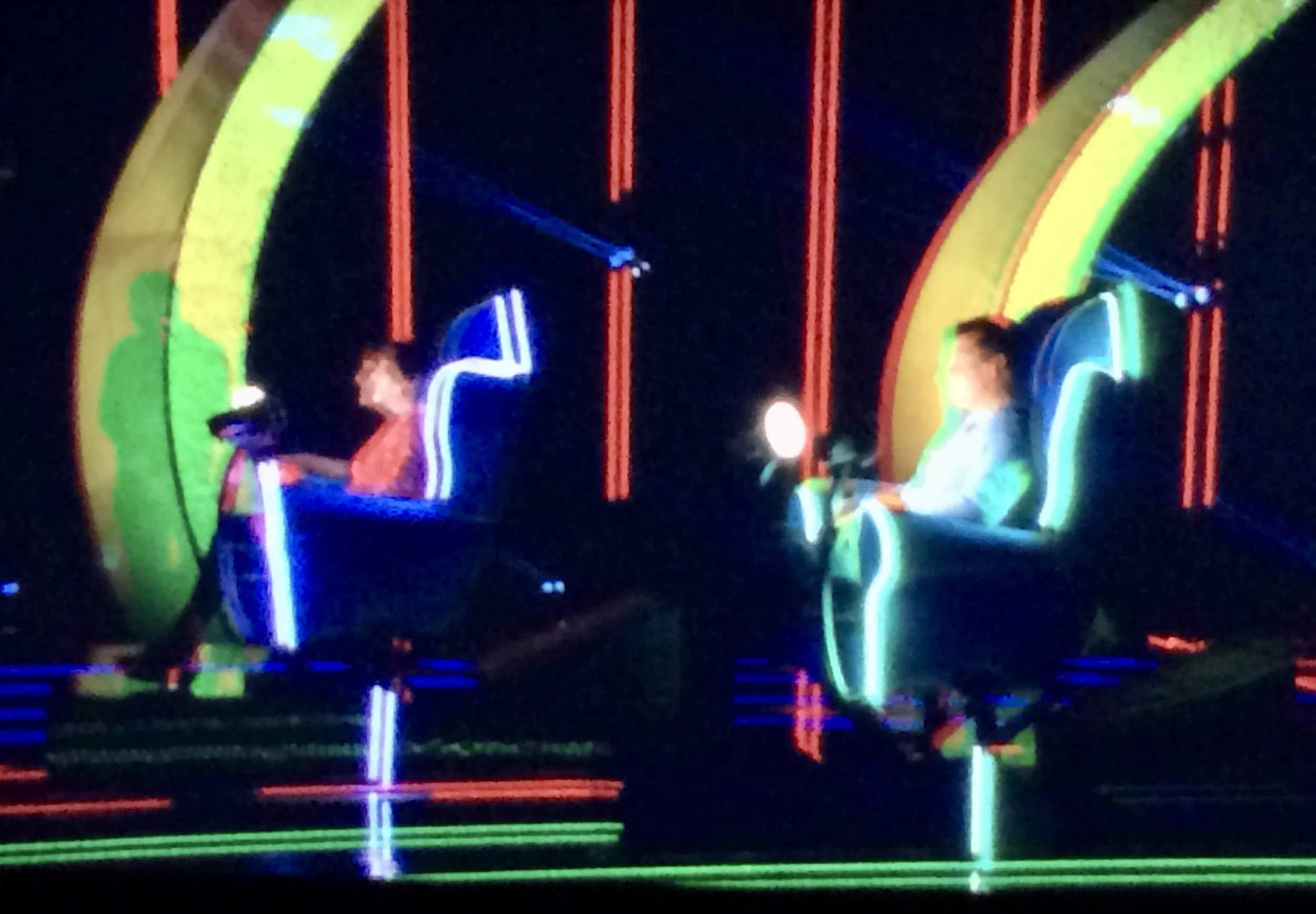 Two people sitting in futuristic glowing chairs with neon outlines on a dark stage.