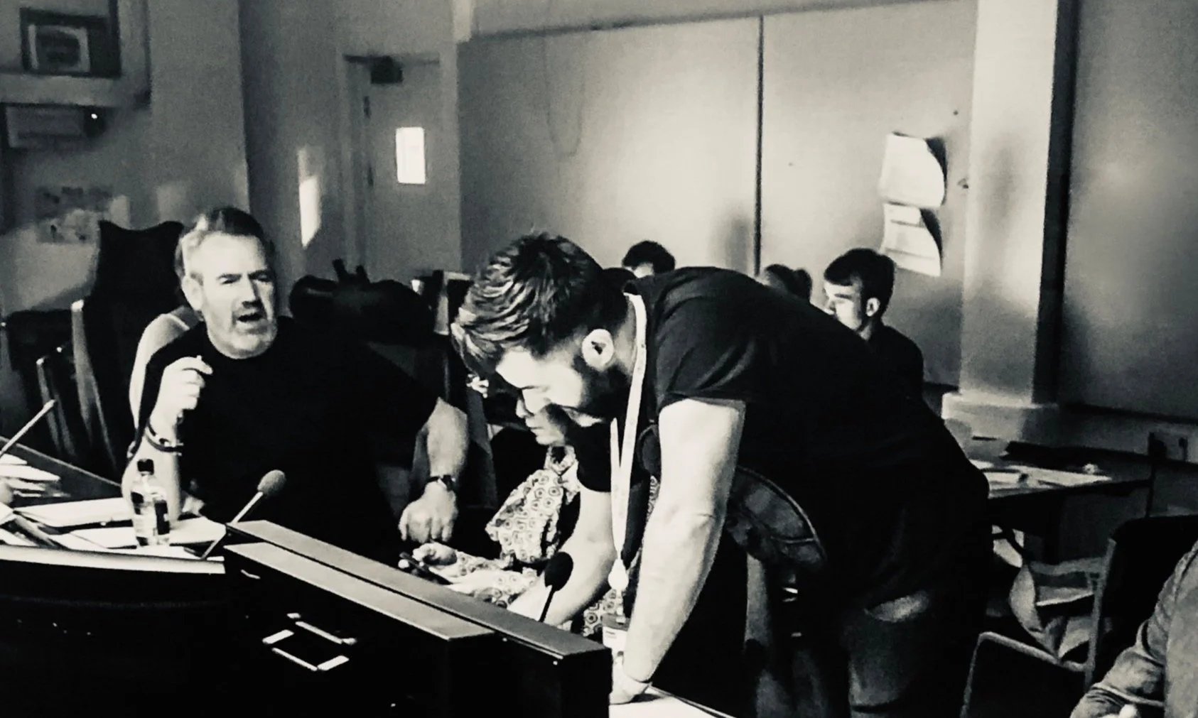 Black and white photo of a group of people in a classroom or office setting, focused on a project or discussion. One person standing over a desk, others sitting with papers and microphones visible.
