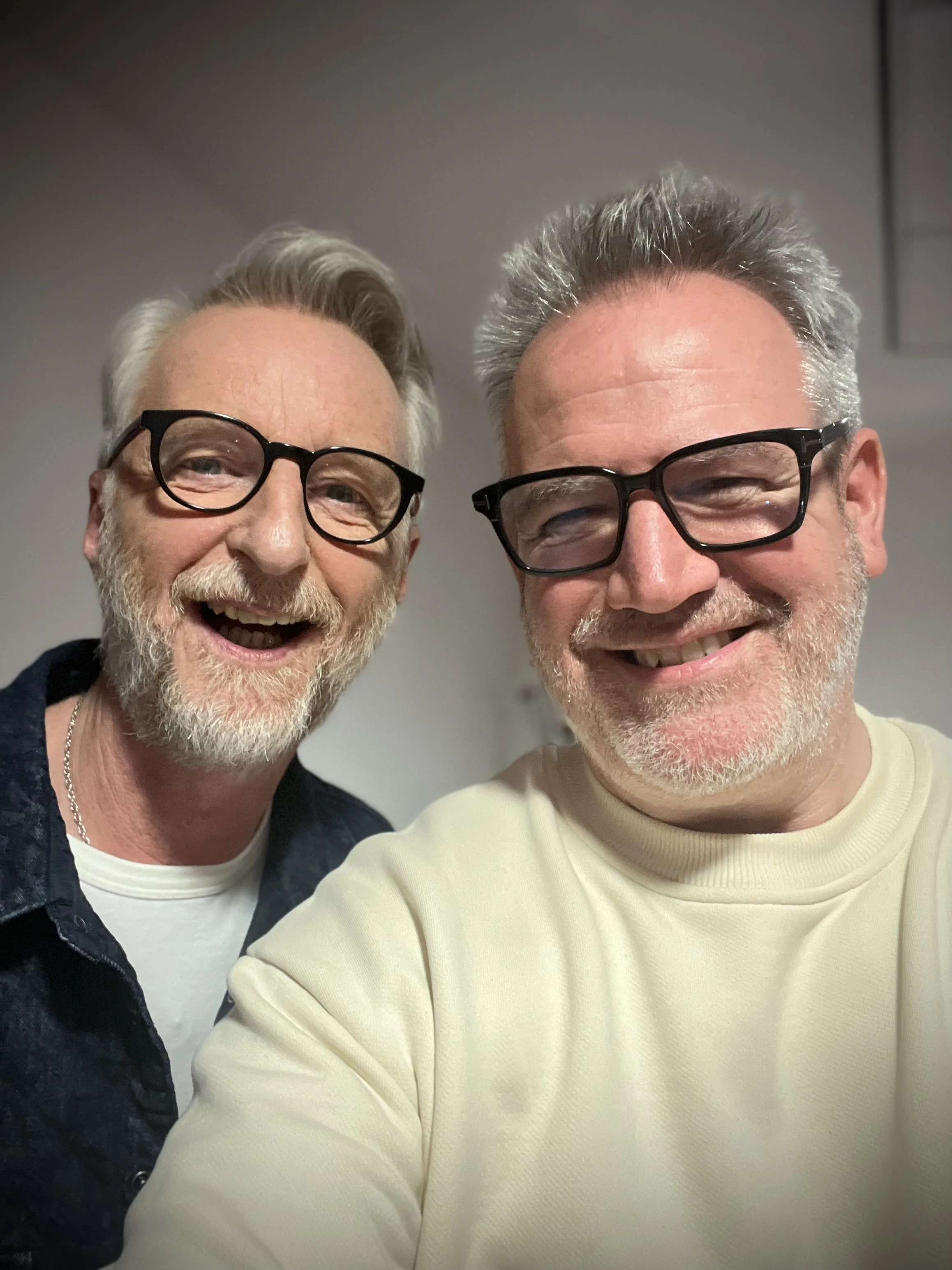 Two smiling men wearing glasses, one in a dark shirt and the other in a light sweater, posing for a selfie in an indoor setting.