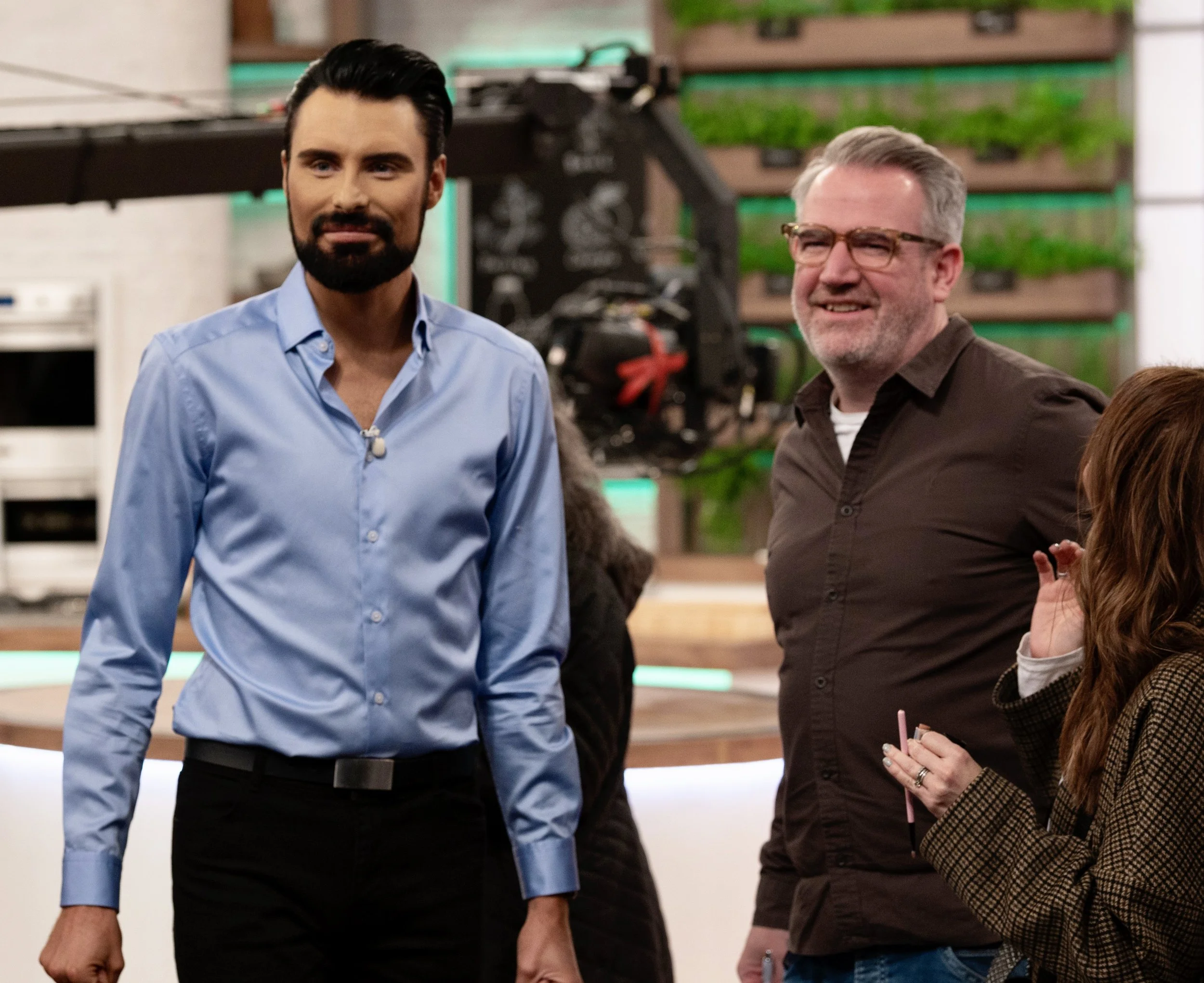 Group of people in a studio setting, including a man in a blue shirt, another man wearing glasses and a dark shirt, and a woman partially visible holding a pen.