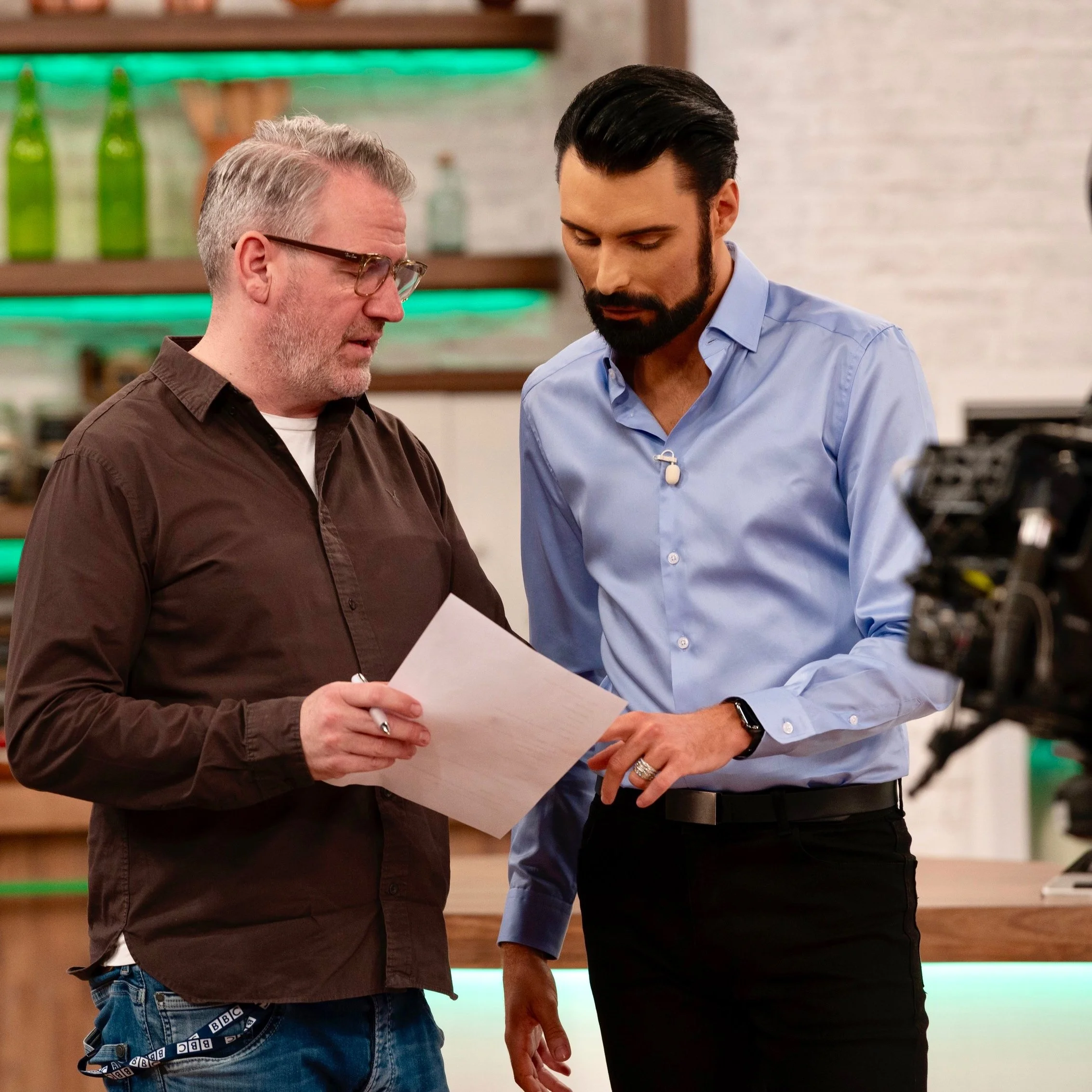 Two individuals in a kitchen or studio setting discussing a document; one is wearing a blue shirt and the other a brown shirt. Shelves with green bottles and kitchen items are in the background.
