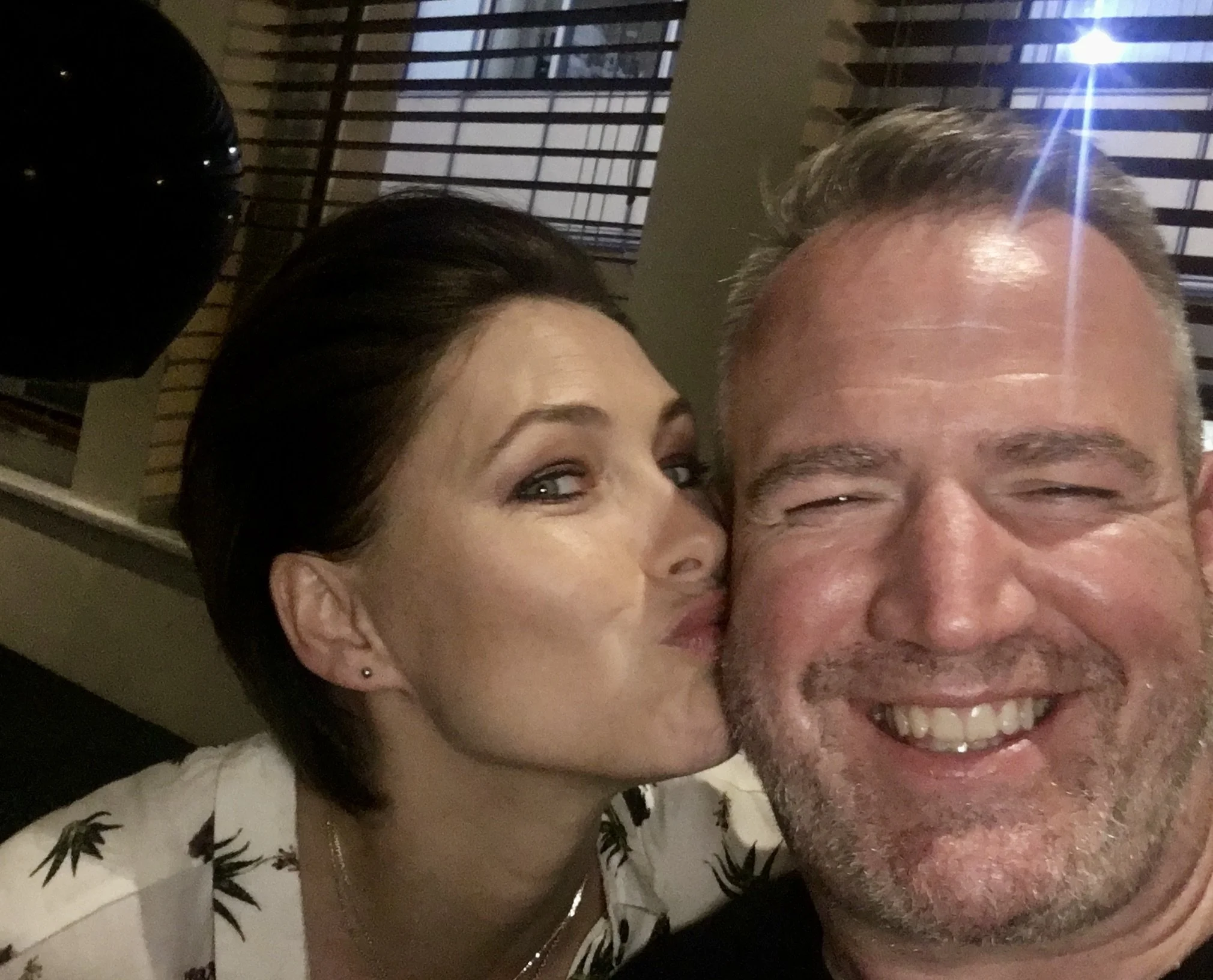 A woman kissing a smiling man on the cheek indoors, with blinds in the background.