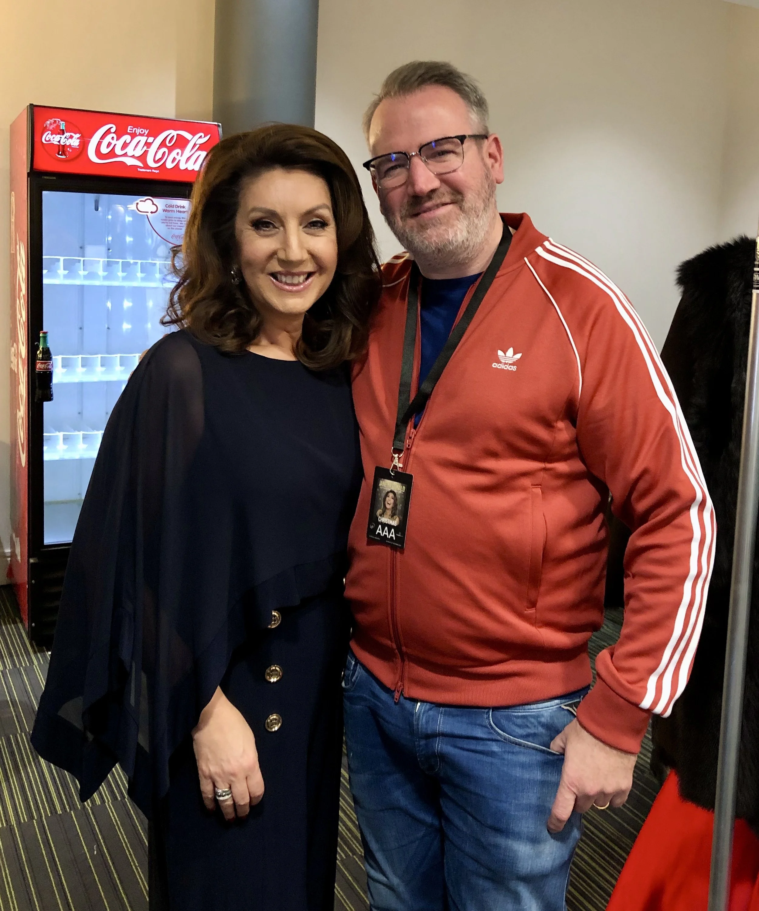 Two people standing together, one in a blue outfit and the other in a red Adidas jacket, with a Coca-Cola refrigerator in the background.