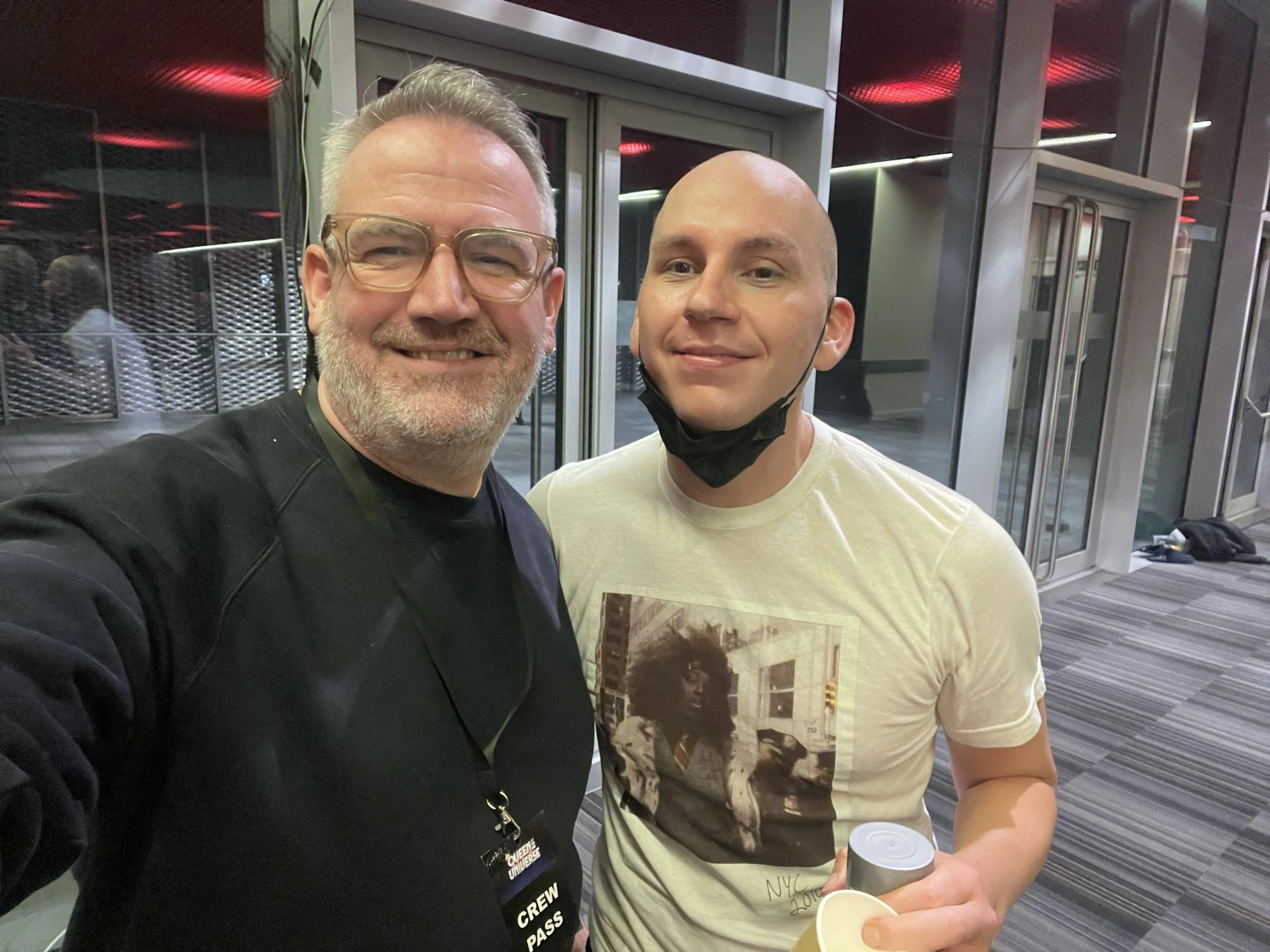 Two people taking a selfie indoors, one wearing glasses and a black crew pass and the other in a white graphic T-shirt holding a drink.