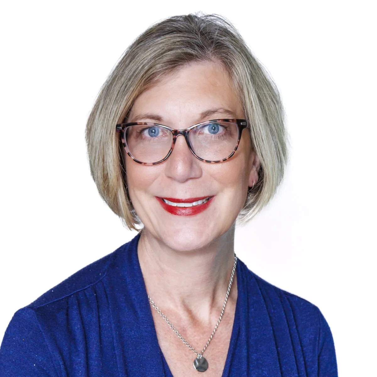 A smiling middle-aged woman with short gray hair, wearing tortoiseshell glasses, a blue top, and a silver necklace with a round pendant, against a white background.