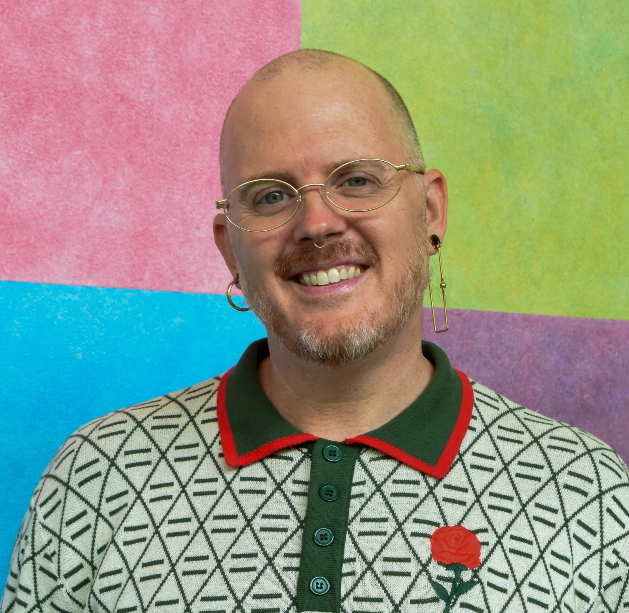 A man wearing glasses, a pink patterned shirt, a pink hat, and pink earrings.