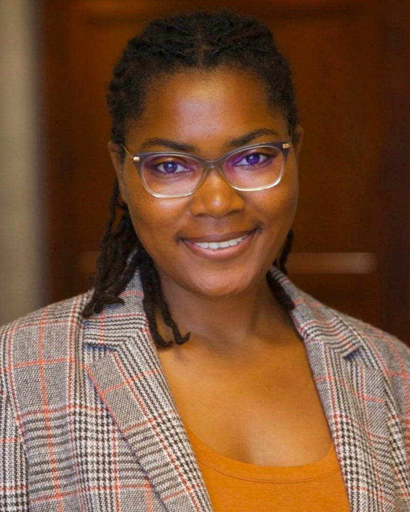 A young African American woman with glasses, wearing a plaid blazer over an orange top, smiling at the camera.