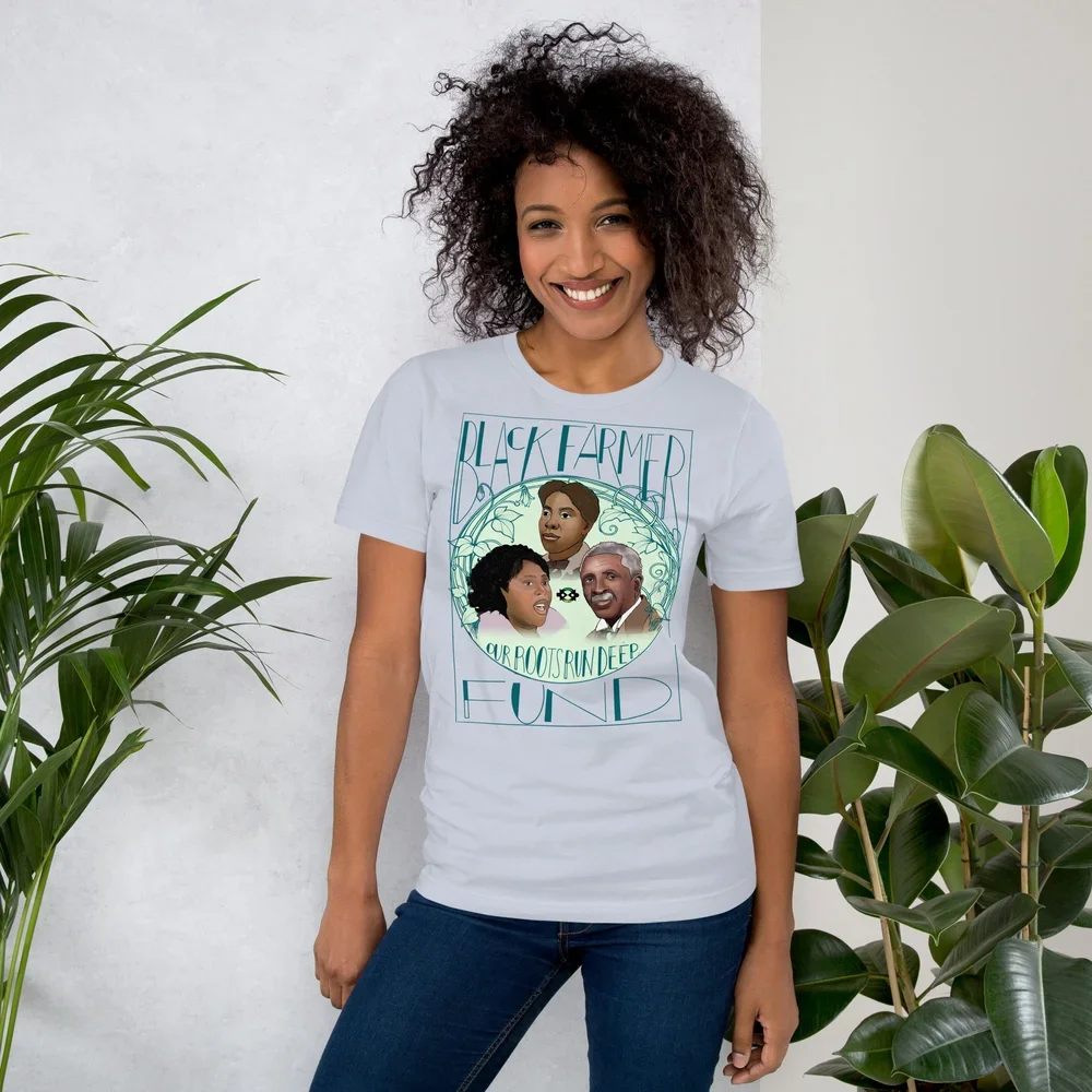 A woman with curly hair smiling and standing next to green plants, wearing a white T-shirt with illustrations of Black figures and text related to Black farmers and roots.