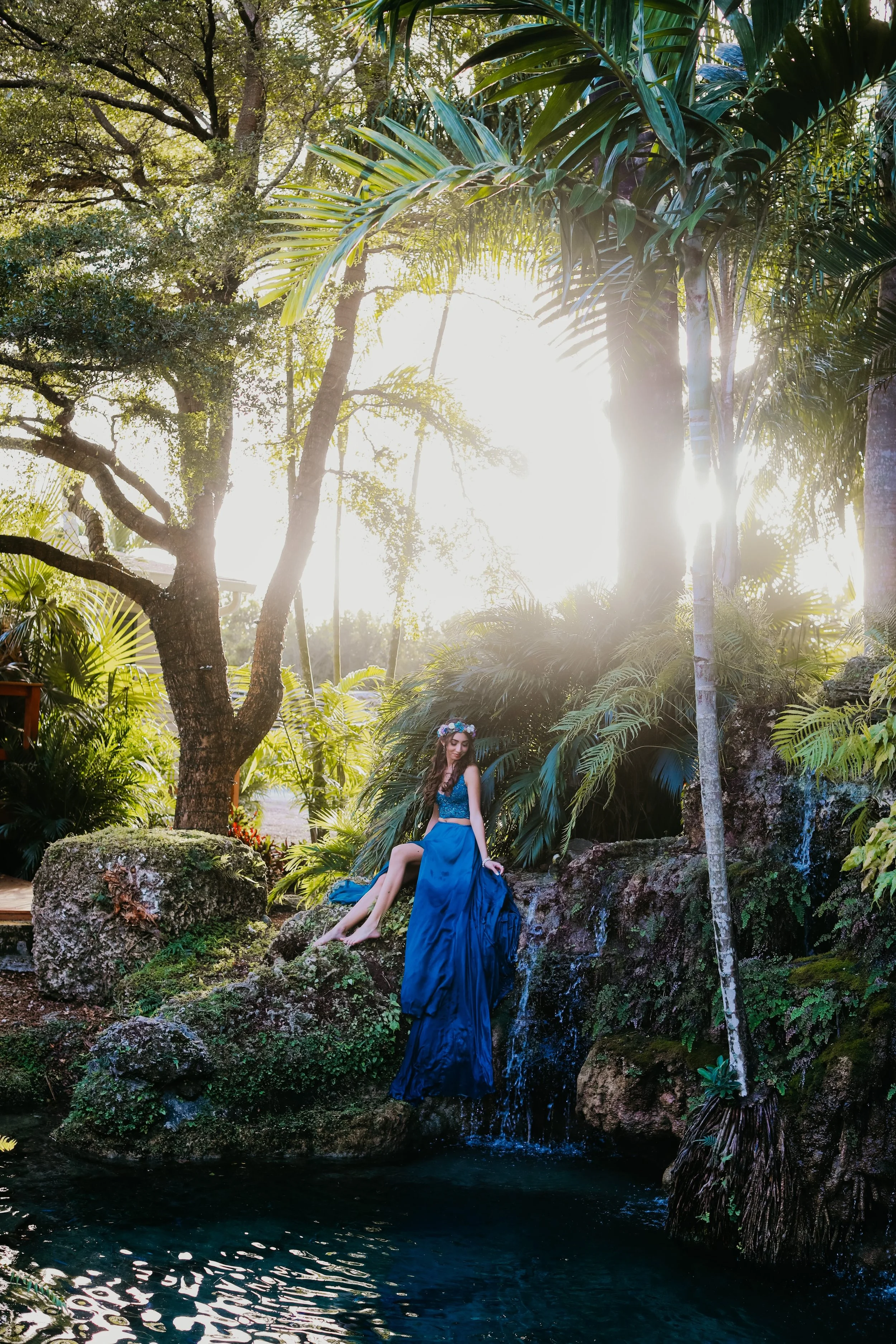 A girl in a blue dress sitting inside a large floral hoop decorated with blue and white flowers over a pond.
