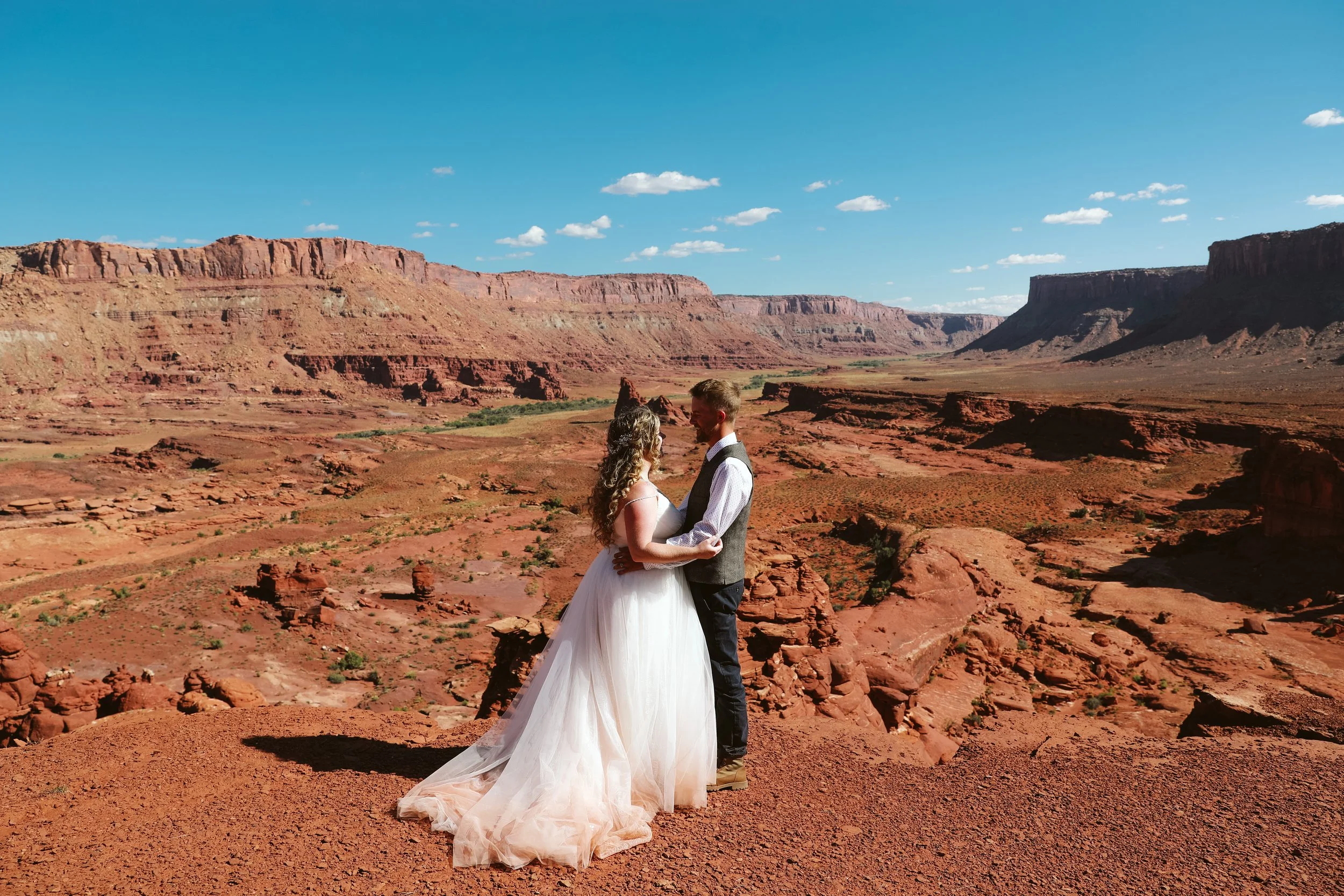 A newlywed couple stands close together in a desert landscape with red rock formations and blue sky in the background.