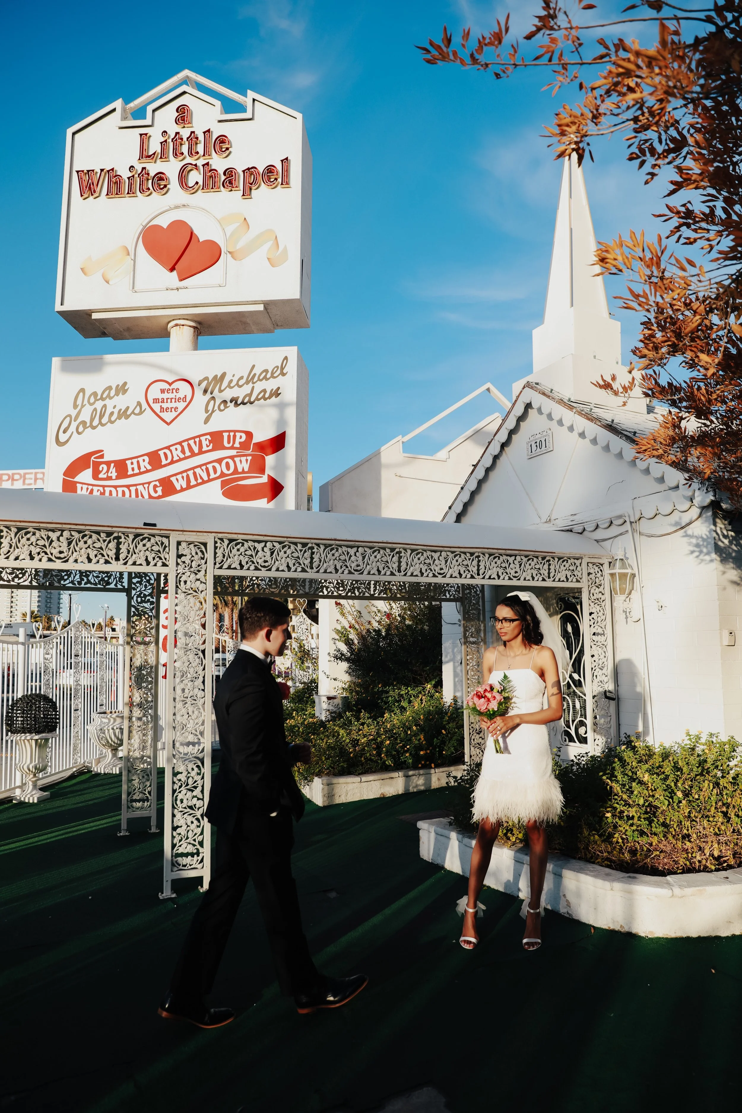A bride and groom stand outside a white chapel with decorative trim. The bride is holding a bouquet of pink flowers, and both are dressed in wedding attire. There are signs above the chapel, one reads 'a Little White Chapel' with a heart symbol, and another indicates a drive-up wedding window. The scene is set on a clear day with a blue sky.