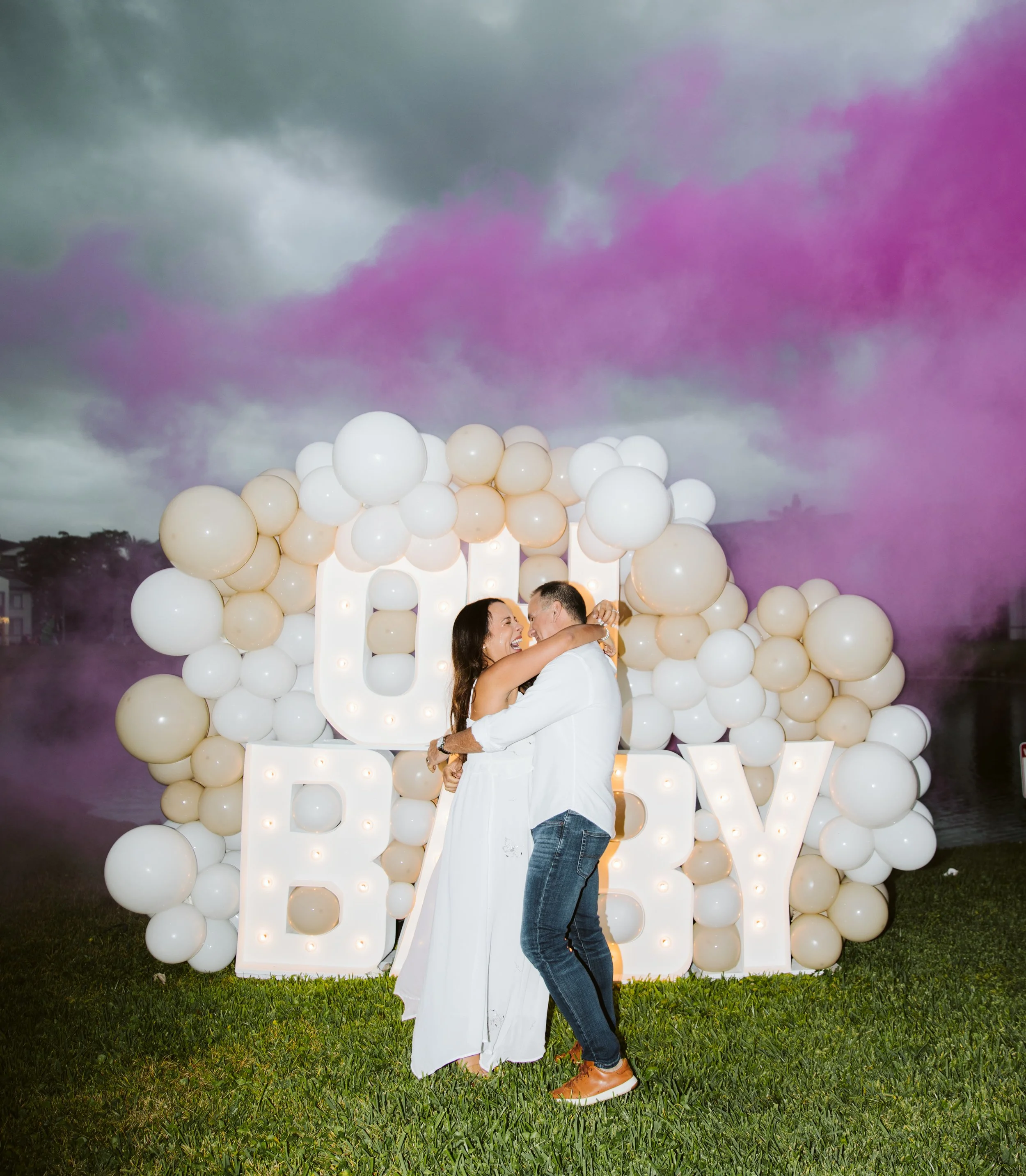 A couple dancing outdoors at a celebration, with a large illuminated 'LOVE' sign decorated with white and beige balloons and purple smoke in the background.