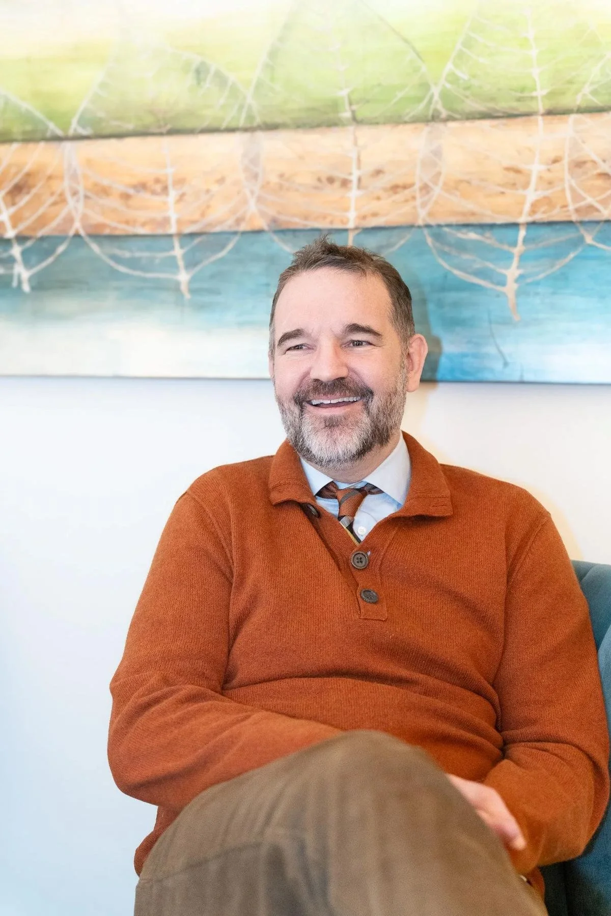 A smiling therapist with a beard and mustache, wearing an orange sweater, a dress shirt, and a patterned tie, sitting indoors in front of a colorful abstract painting.