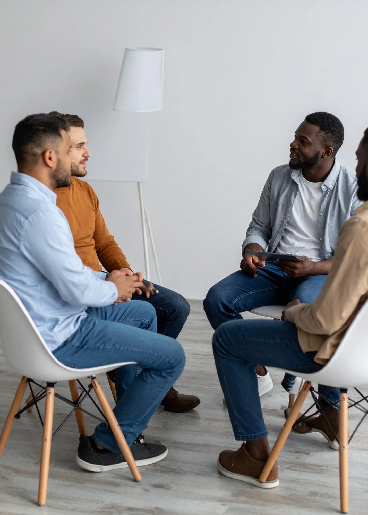 A group of five diverse men sitting in a porn support group, engaging in a discussion in a bright, minimalistic room with white walls and a floor lamp.