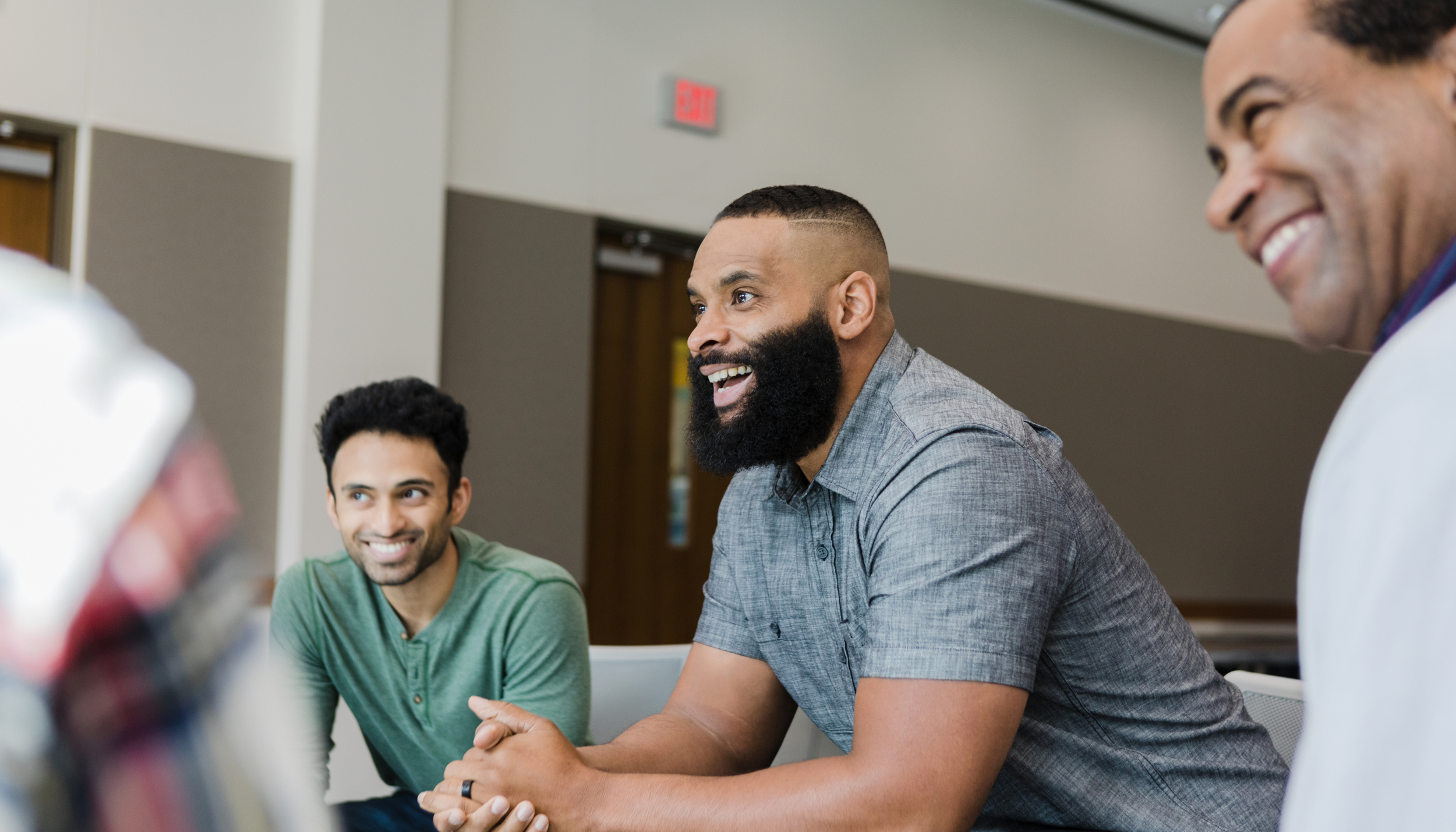 Three men sitting at a table and talking, smiling, in a casual indoor setting.