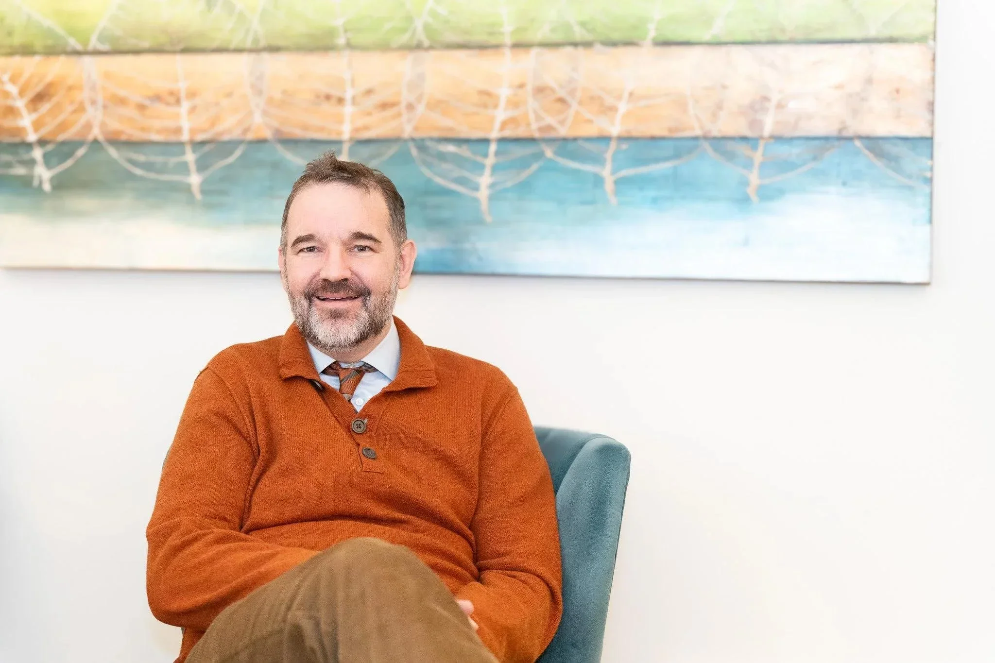 A male therapist with a beard and short hair, wearing an orange sweater over a collared shirt and tie, sitting in a turquoise chair in front of a colorful painting with abstract patterns.