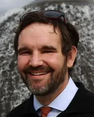 A male addiction therapist with dark hair and a beard smiling outdoors in Seattle, with a rocky background.