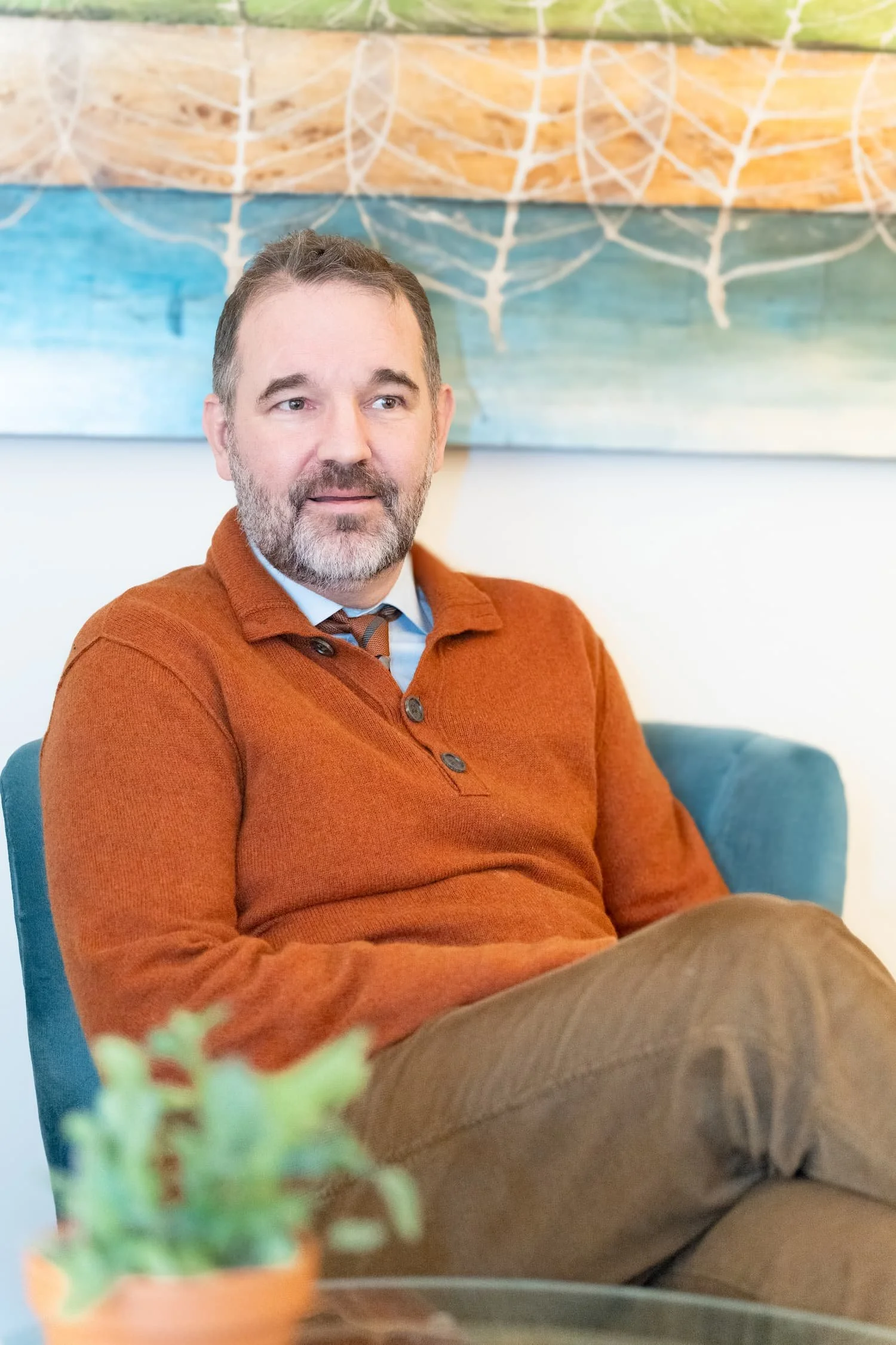 A therapist with a beard, wearing an orange sweater over a collared shirt and tie, sitting on a blue couch in front of a colorful abstract wall art. He appears to be engaged or in thought.