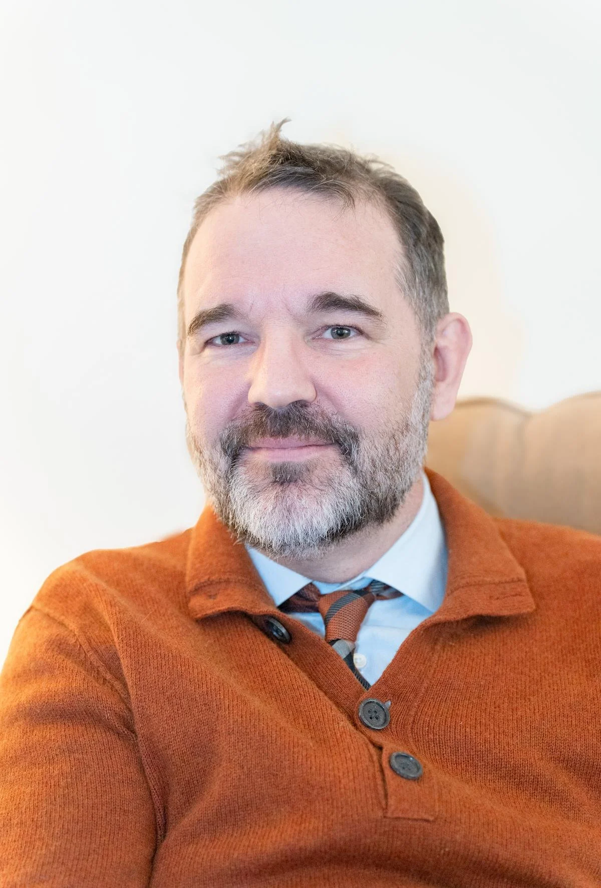 A male therapist with a beard and gray hair, wearing a rust-colored sweater, white shirt, and a striped tie, sitting in a wooden chair against a plain white background.