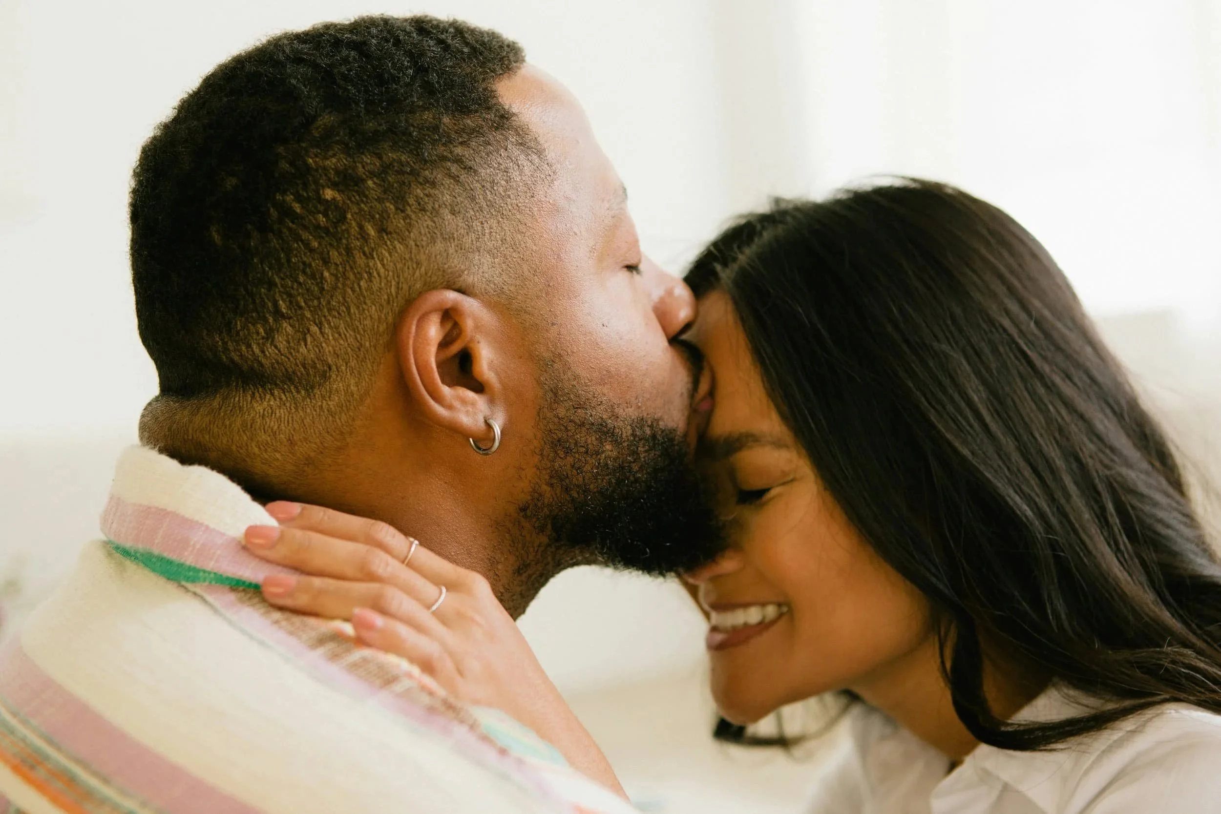 A man and woman sharing an intimate moment, the man is kissing the woman on her forehead and she is smiling with closed eyes.