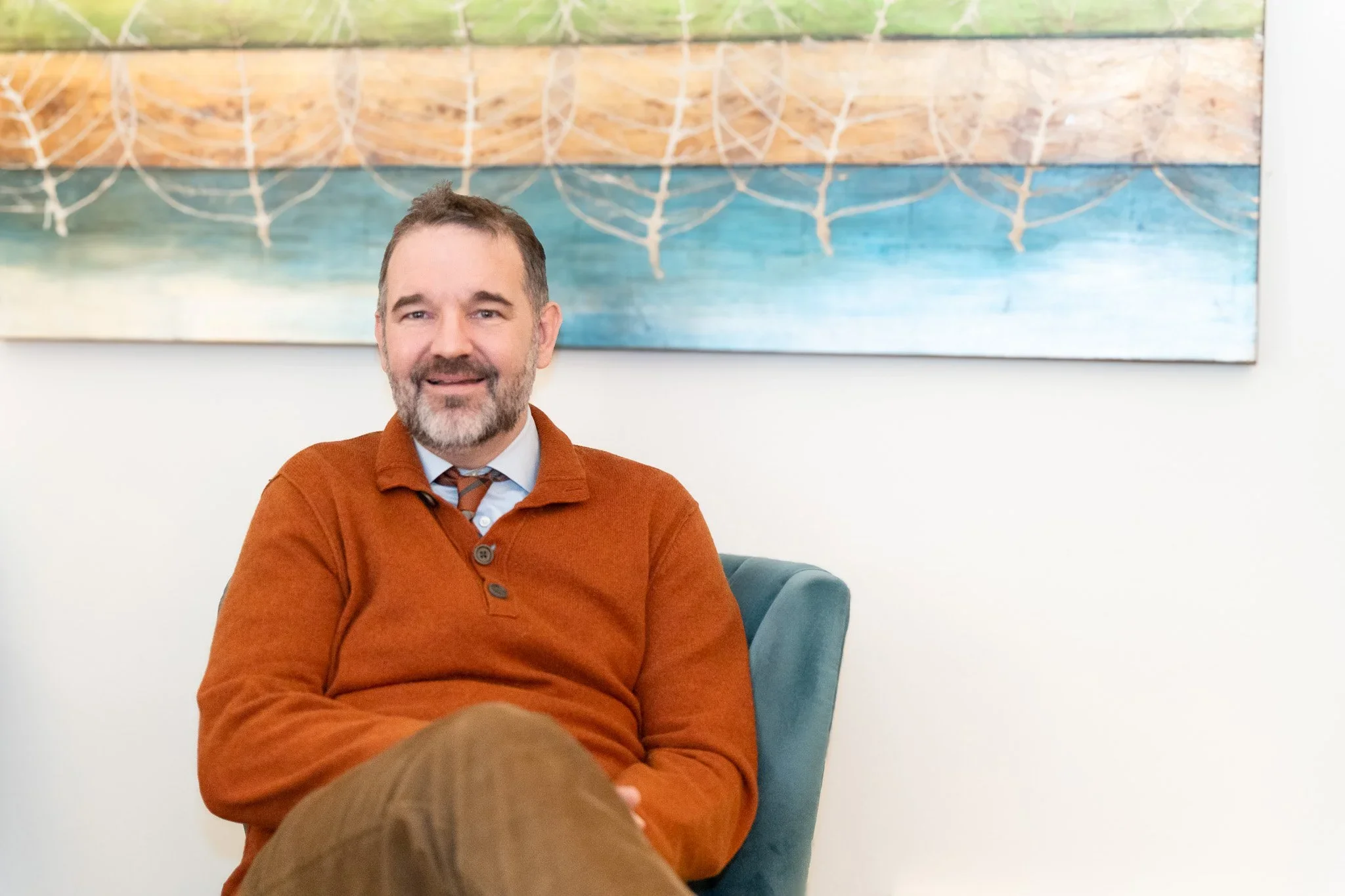 A therapist with a beard and mustache, wearing a rust-colored sweater, sits on a blue chair in front of a colorful abstract painting, smiling at the camera.