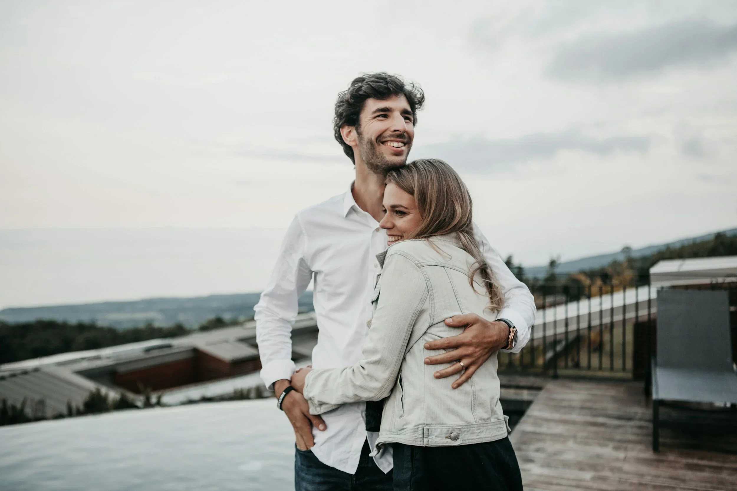 A man and woman hugging outdoors near a railing with cloudy sky and mountains in the background.